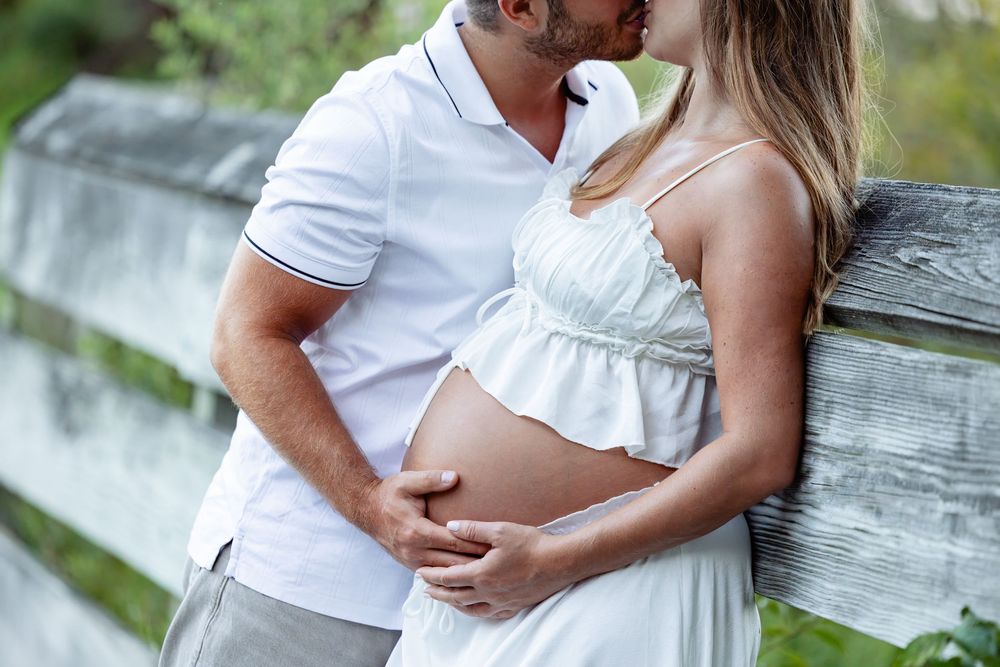 Couple sharing a tender kiss outdoors, with pregnant woman in white dress cradling her baby bump by a wooden fence.