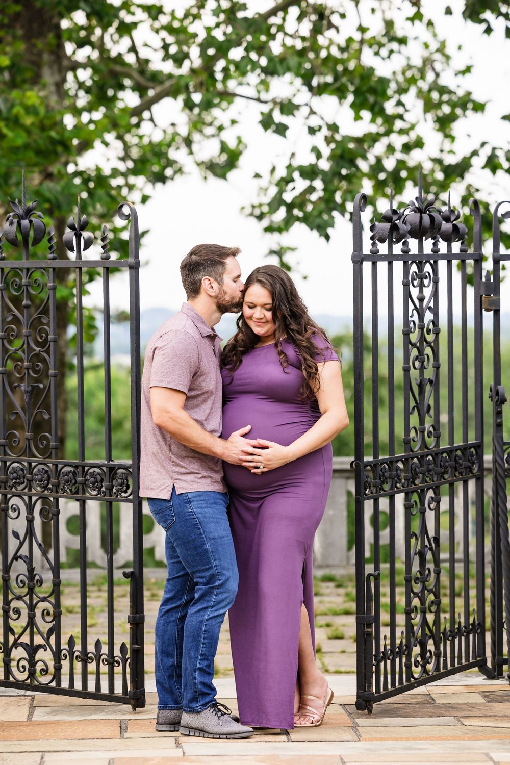 Couple sharing a tender maternity moment by ornate iron gates, woman in purple dress cradling baby bump.