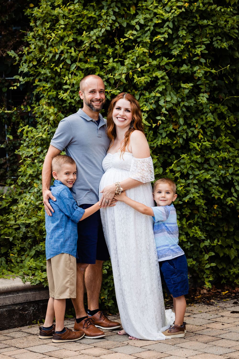 Pregnant woman in white lace gown poses with husband and two young sons in front of lush green hedge.