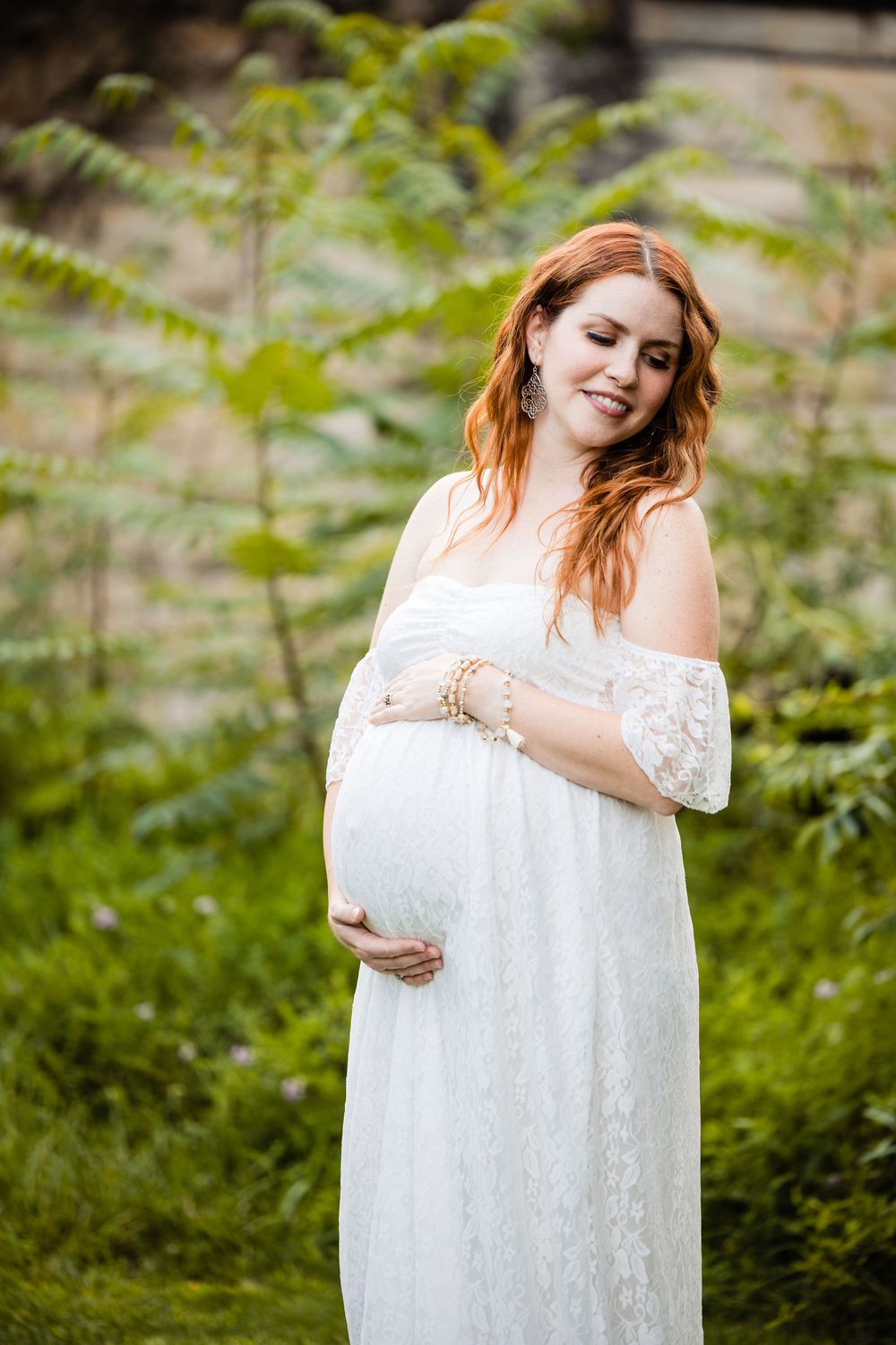 Pregnant woman in white lace off-shoulder dress cradling baby bump outdoors among lush green ferns.