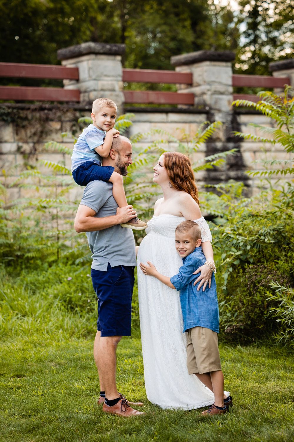 Pregnant woman in white dress with husband and two young boys posing outdoors in lush green garden setting.