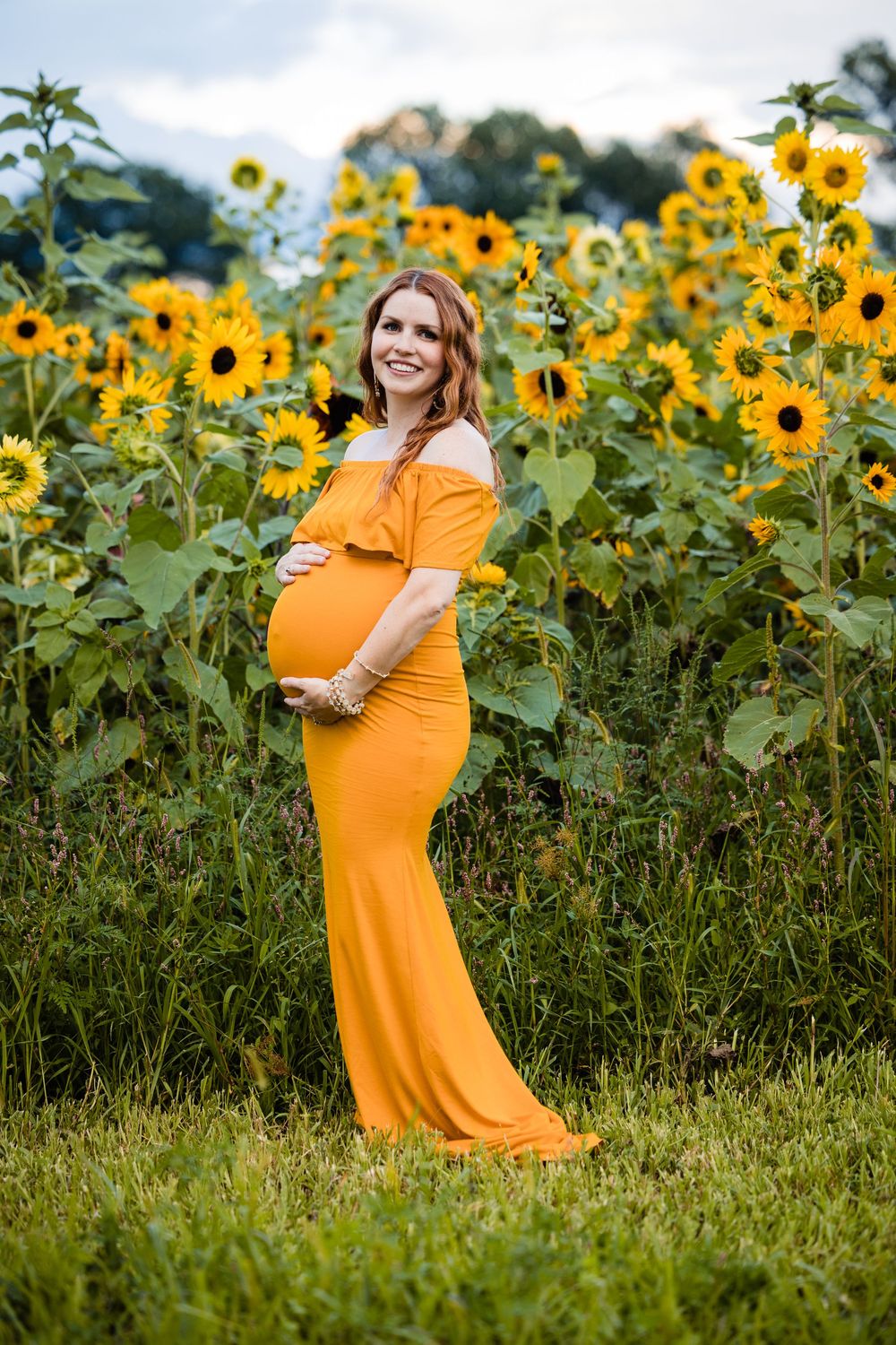 Smiling pregnant woman in yellow off-shoulder gown cradling baby bump in a vibrant sunflower field.