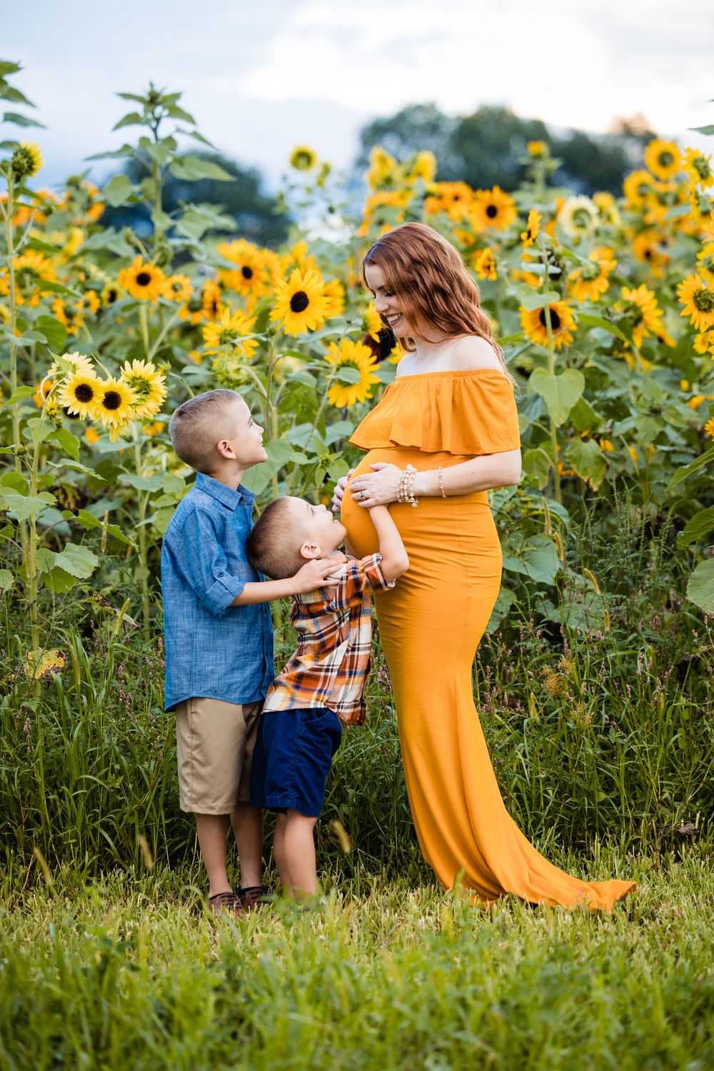 Pregnant woman in orange gown with two young boys touching her belly in a sunflower field.