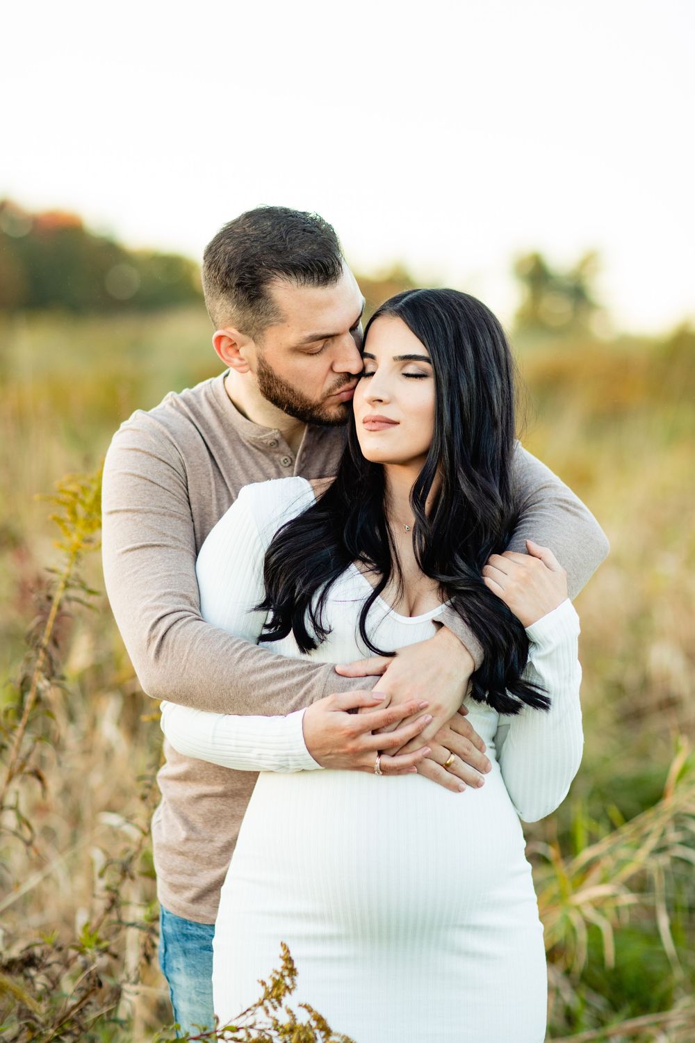 A man tenderly embraces a pregnant woman in a white dress outdoors in a golden field during sunset.