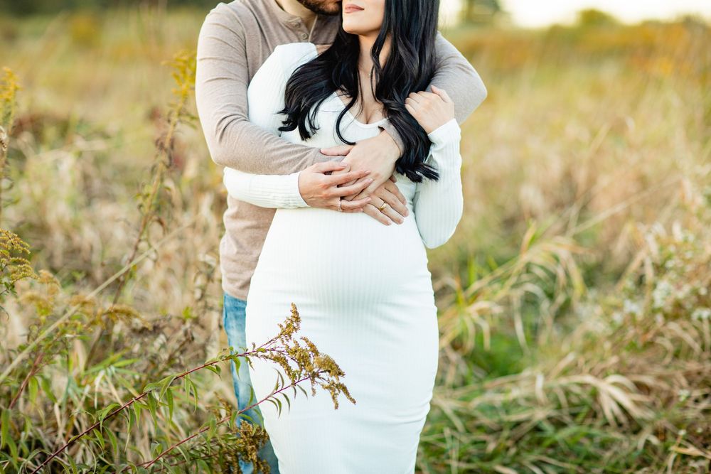 Pregnant woman in white dress embraced by partner in golden field during maternity photo session.