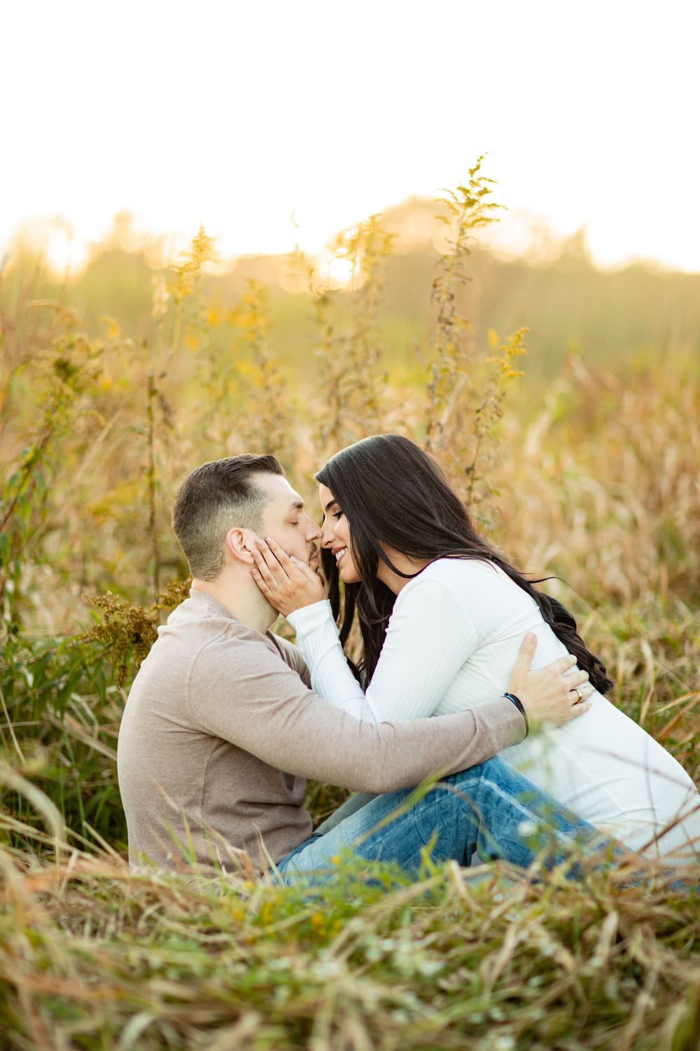 Couple sharing a romantic moment sitting in golden autumn field grass, woman touching mans face tenderly at sunset.