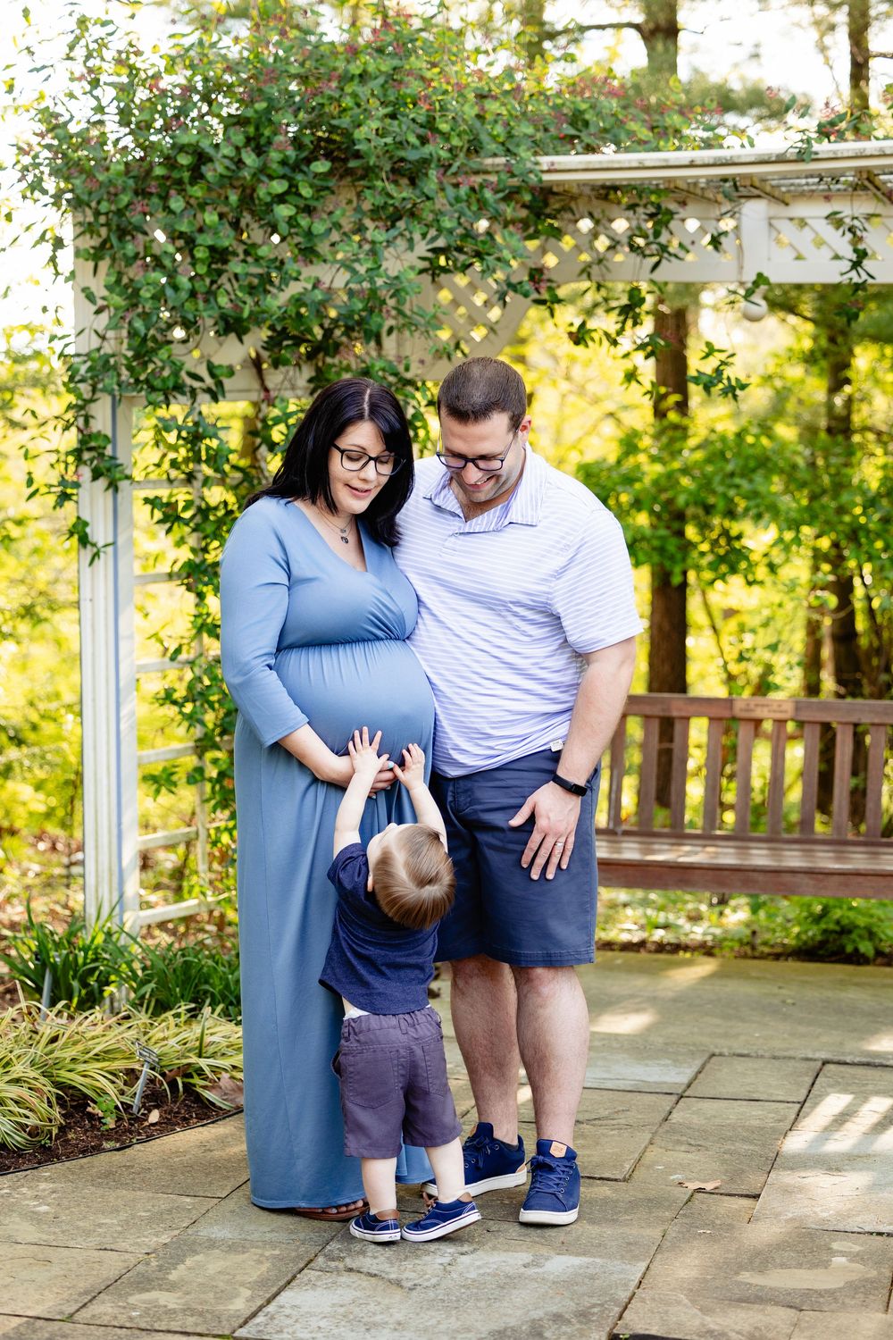 Pregnant woman in blue dress with husband and toddler son touching her baby bump in a garden setting.