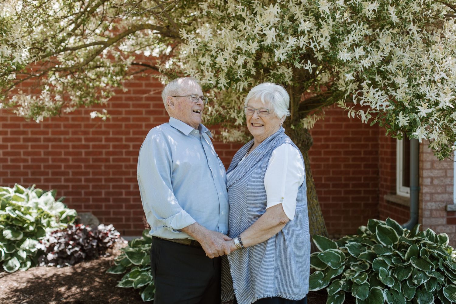 Elderly couple sharing a tender moment together in front of a brick building with blooming trees.