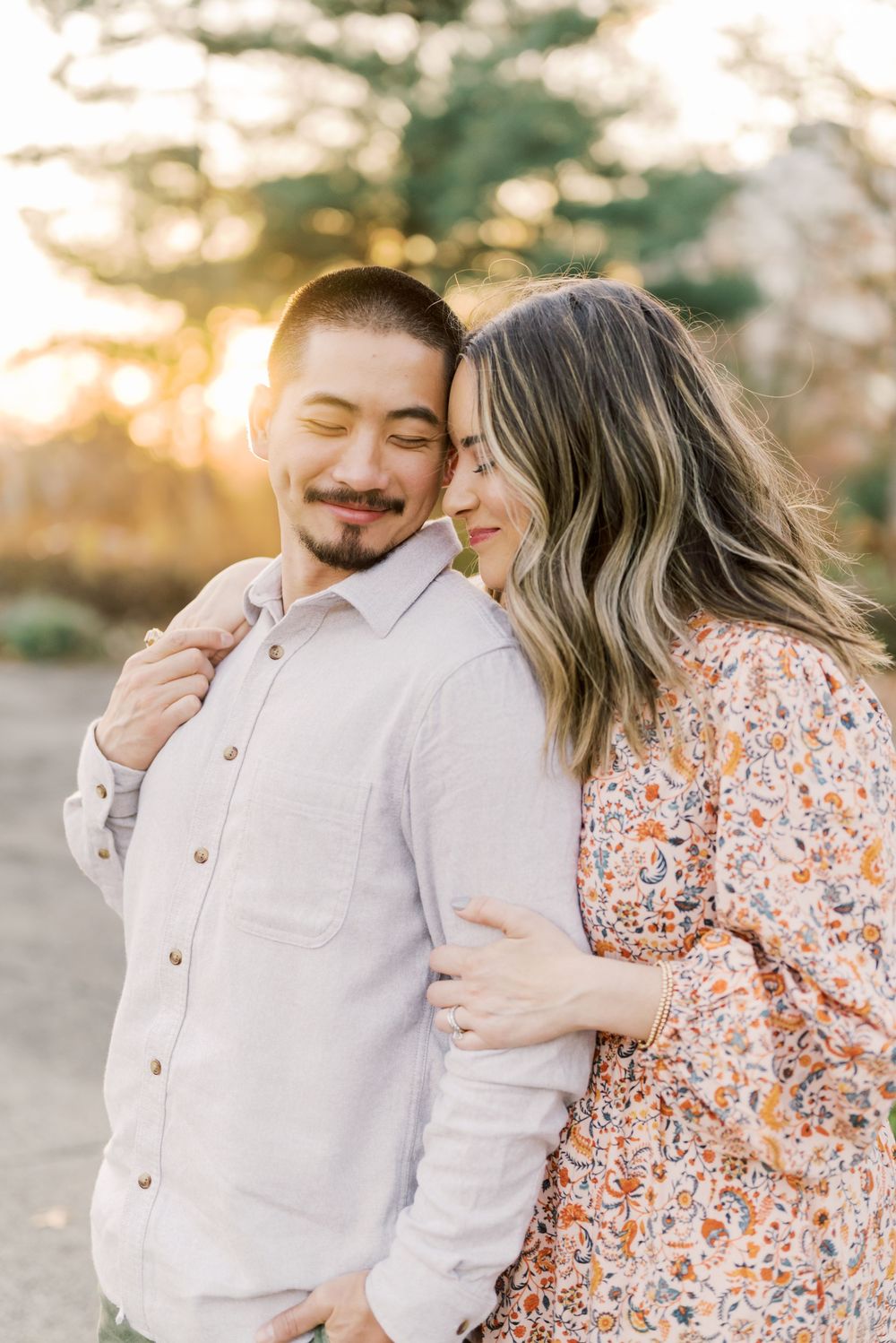A couple embraces warmly at golden hour, the woman in a floral dress resting her head on the man's shoulder outdoors.