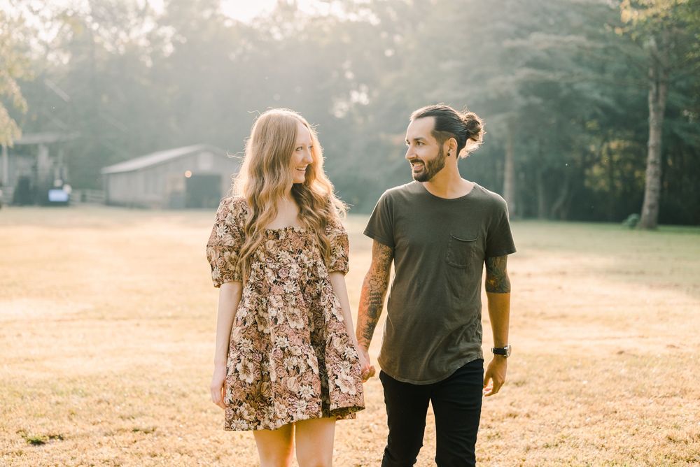 Couple holding hands walking outdoors in golden sunlight, woman in floral dress and man in dark t-shirt.