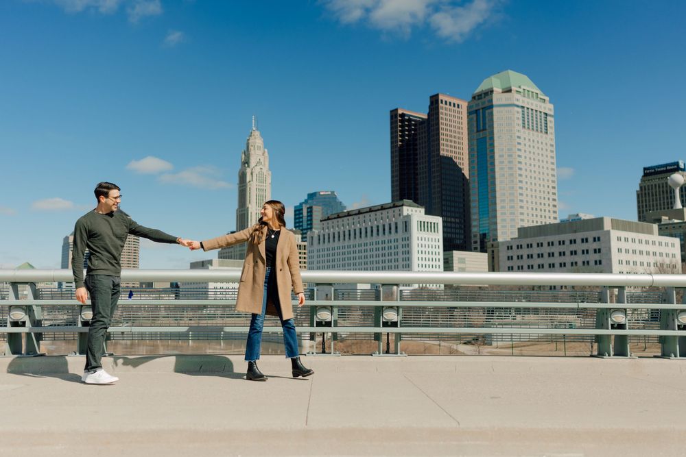 Two figures walking along a riverside promenade with downtown buildings in view.