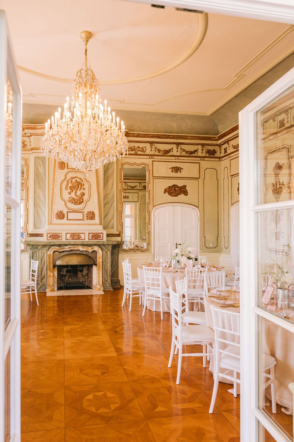 Elegant ballroom with crystal chandelier, white chairs, parquet flooring, and ornate fireplace in classical European style.