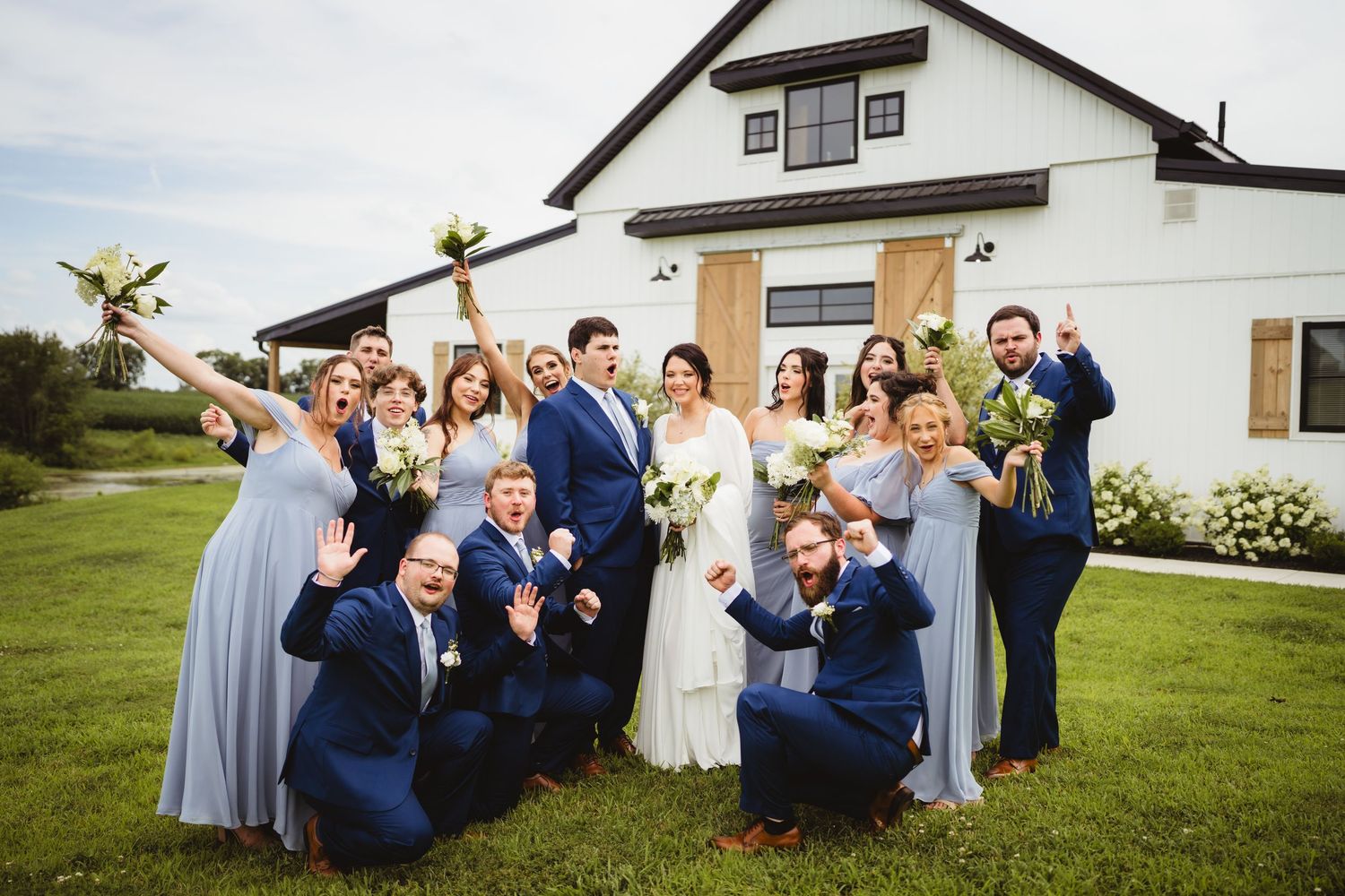 Wedding party members in blue and gray attire pose playfully together on a grassy lawn area in front of homes.