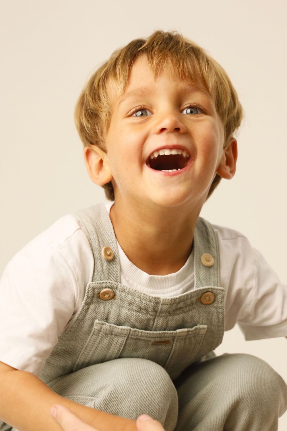A joyful child wearing gray overalls and a white shirt laughs while sitting against a light background.