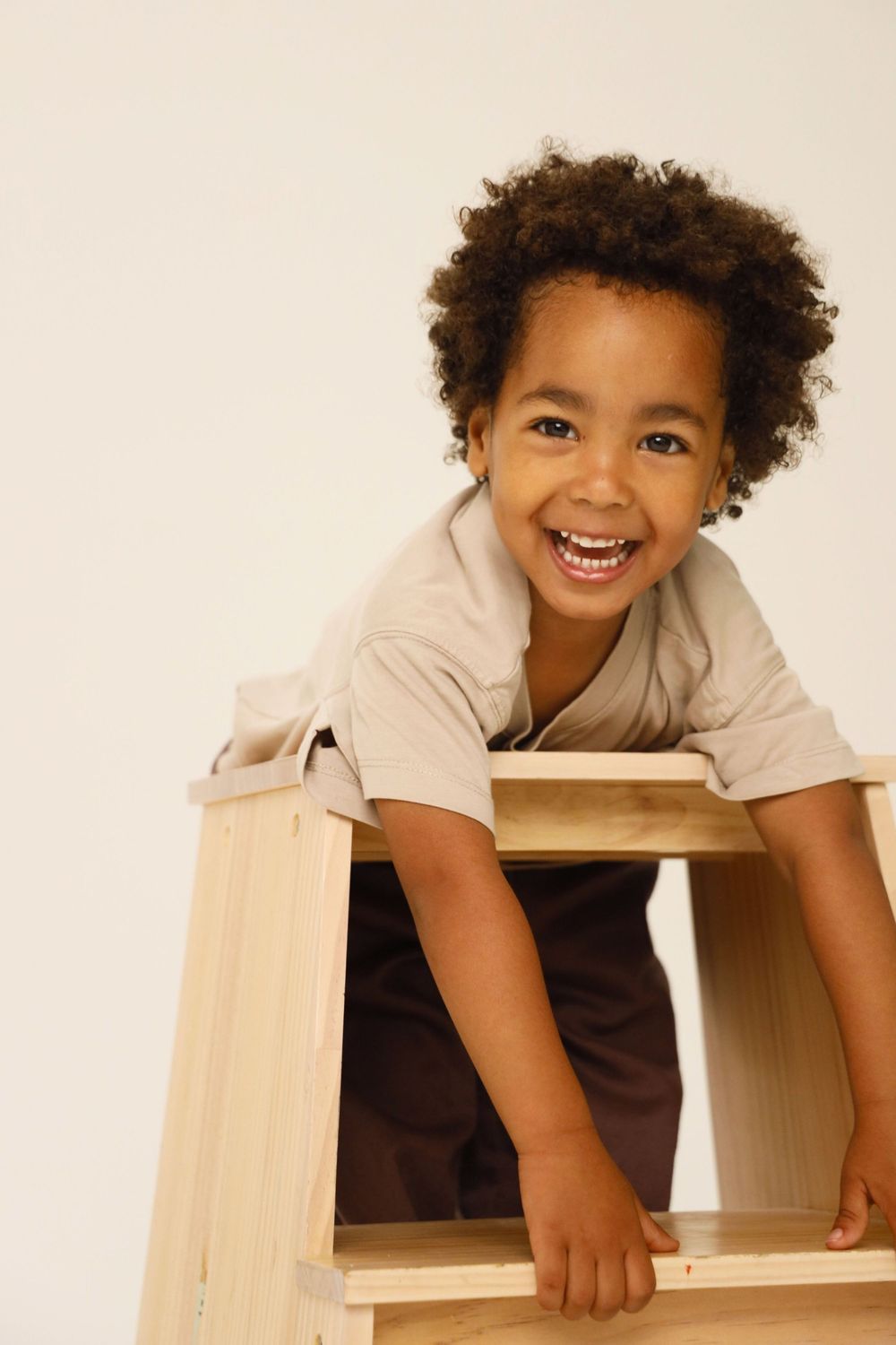 Joyful child with curly hair peeks playfully from behind wooden furniture in a bright setting.