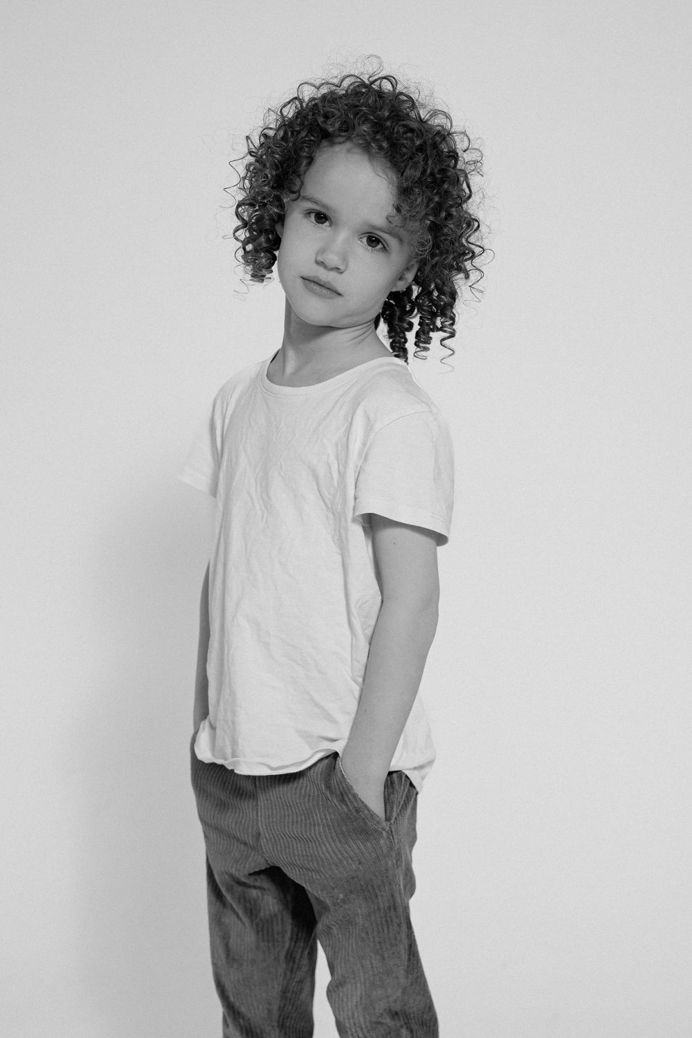 A child with curly hair wearing a white t-shirt and gray pants poses against a light background in a series of photos.