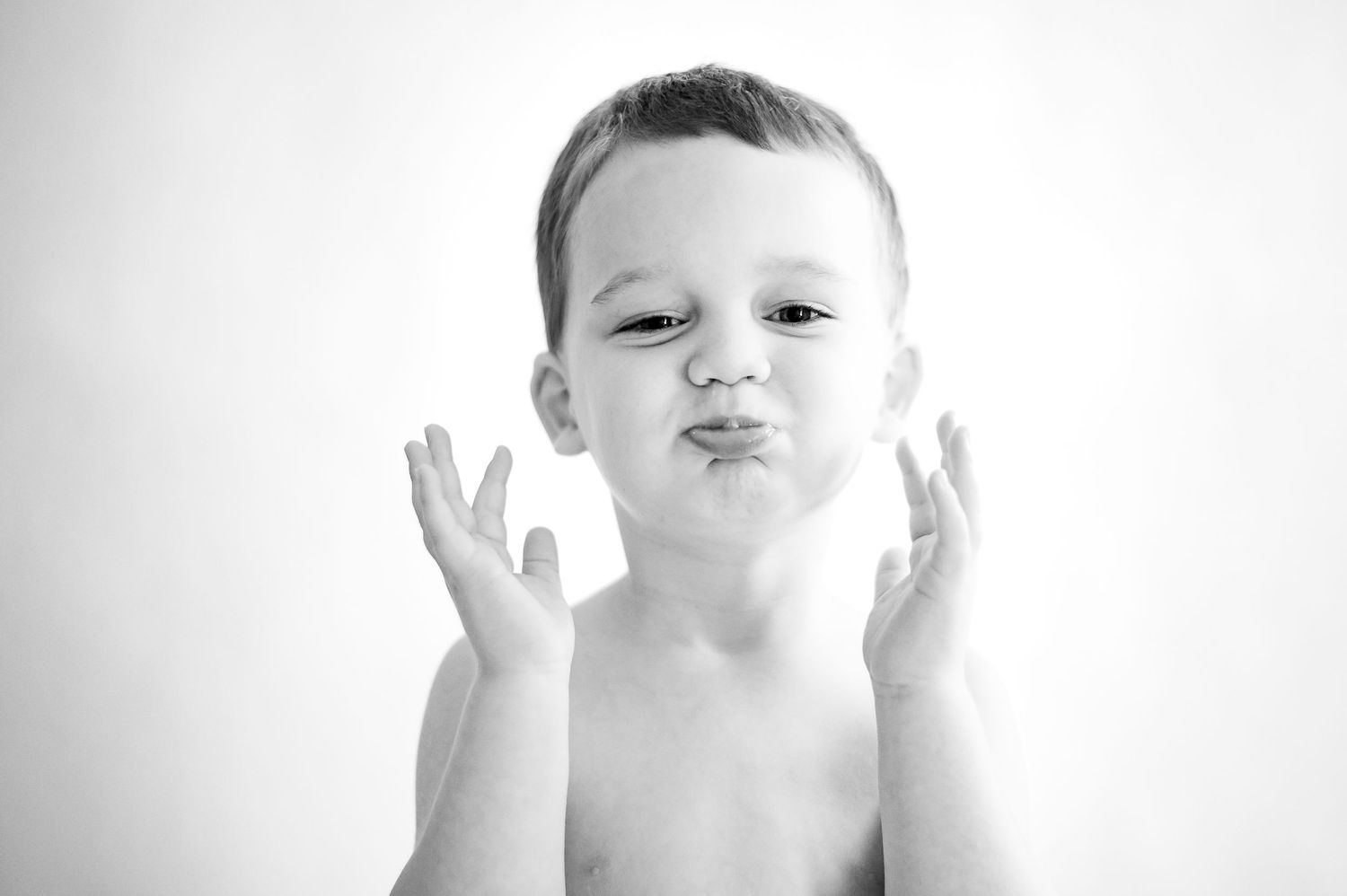 Black and white photo of a child making a playful kissy-face expression with hands up against a light background.