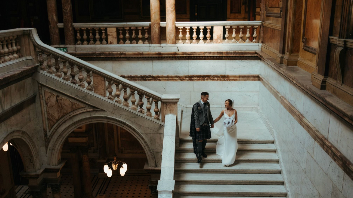 Bride and groom descend ornate marble staircase in grand historic building during elegant wedding ceremony.