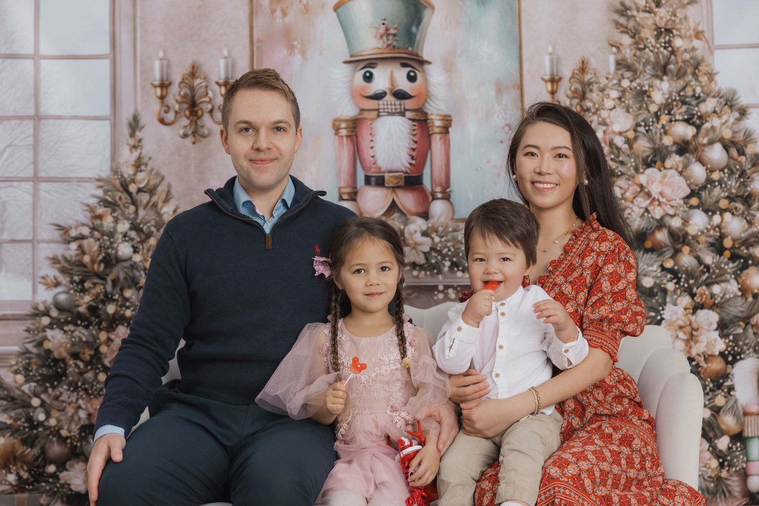 A family sits together in coordinated outfits for holiday photos with decorated Christmas trees in the background.