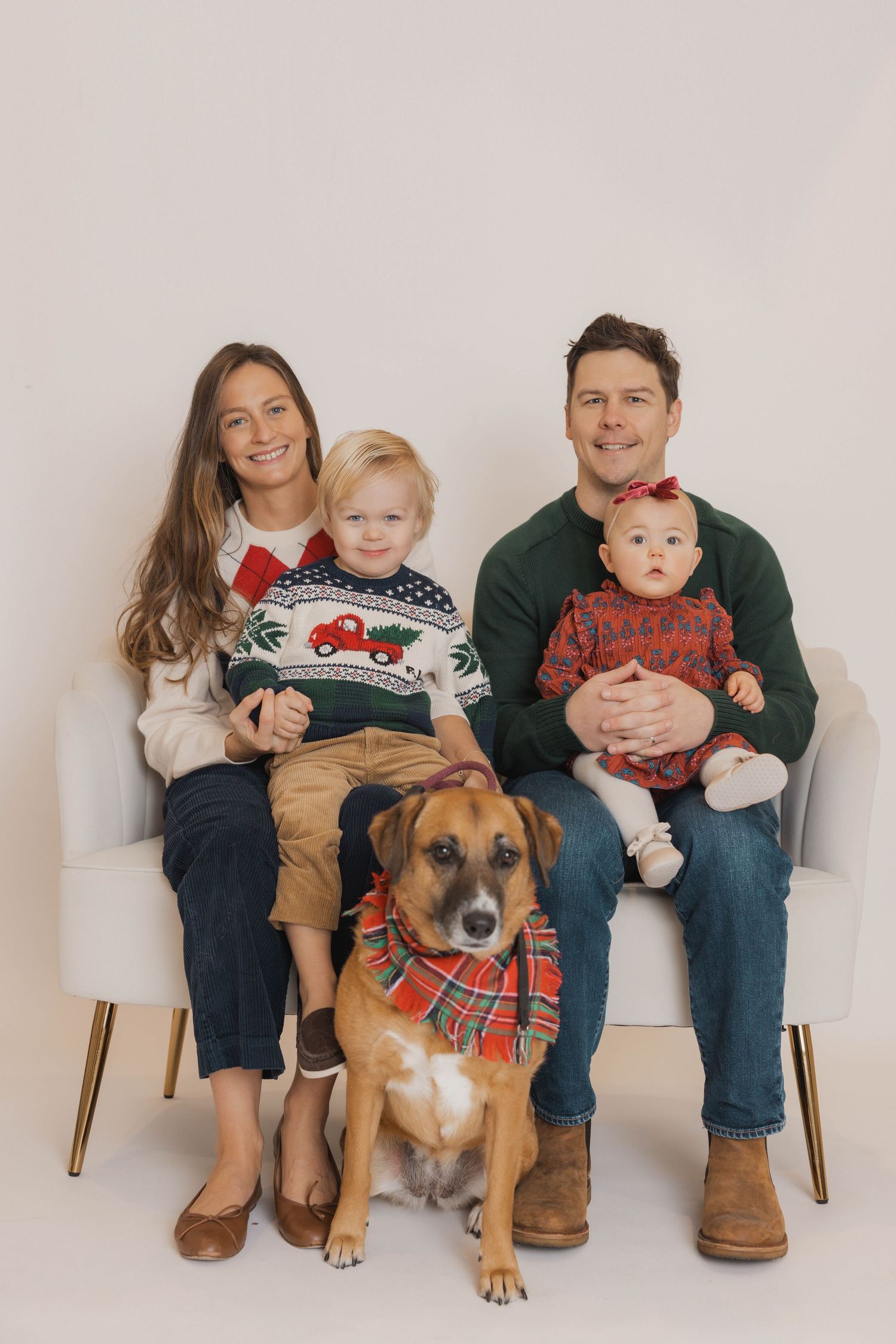 A family and their dog wearing matching holiday sweaters sit together on wooden chairs for a portrait session.