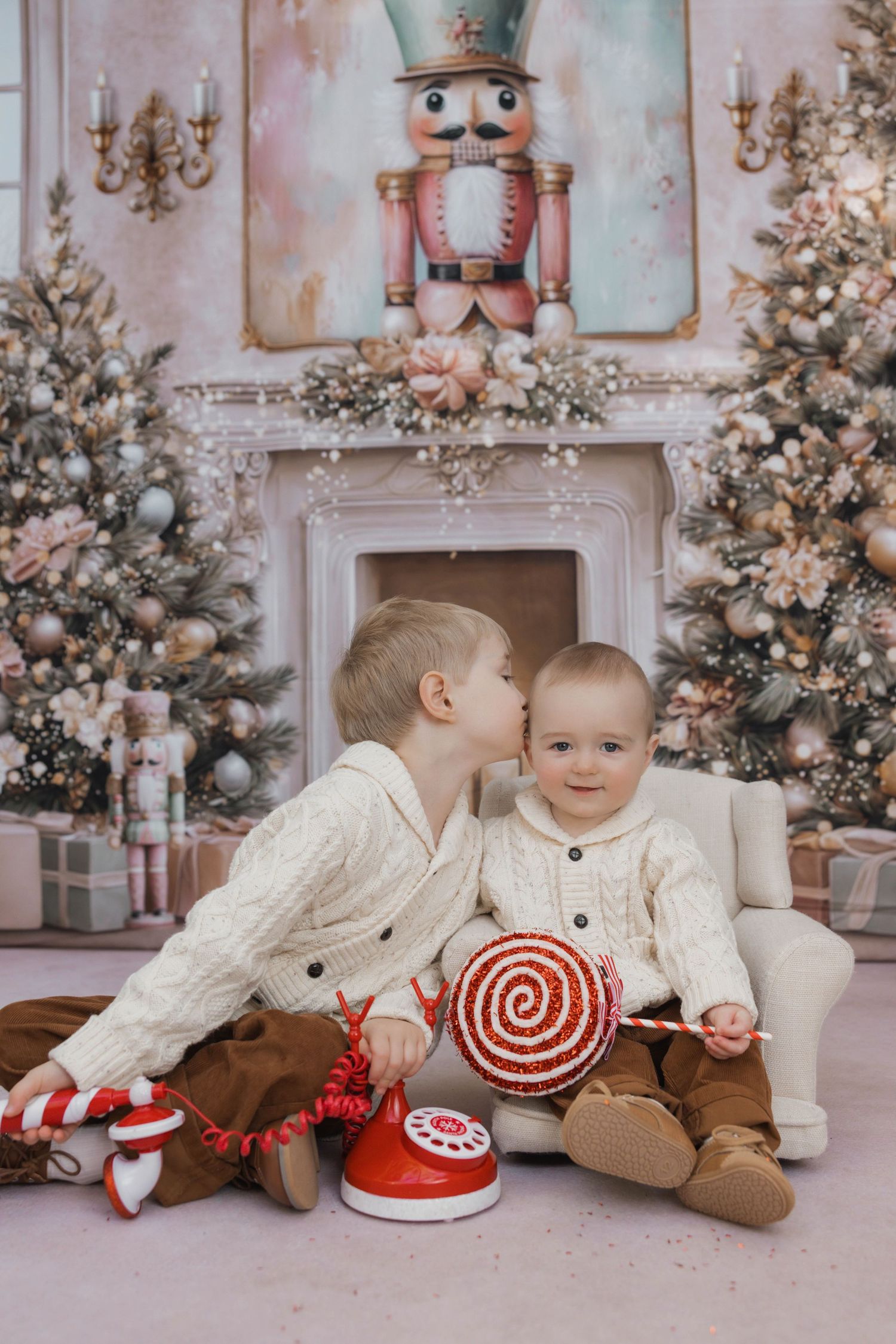 Two toddlers in white sweaters lay on floor beside Christmas trees and nutcracker decorations in festive holiday setting.