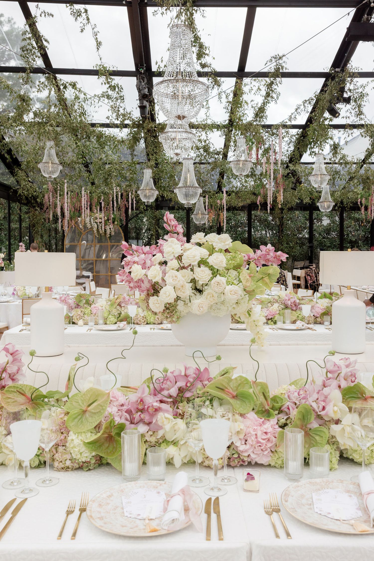 Elegant wedding table setting with pink and white floral arrangements under crystal chandeliers in a glass-ceiling venue.