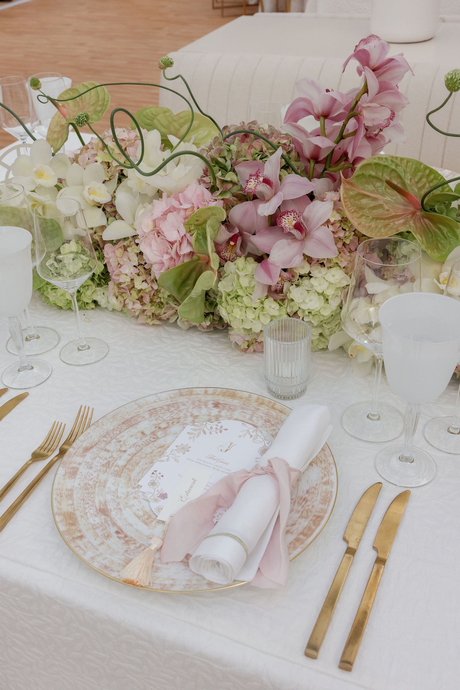 An elegant table setting with pink floral centerpiece, golden cutlery, and vintage patterned plates on white linens.