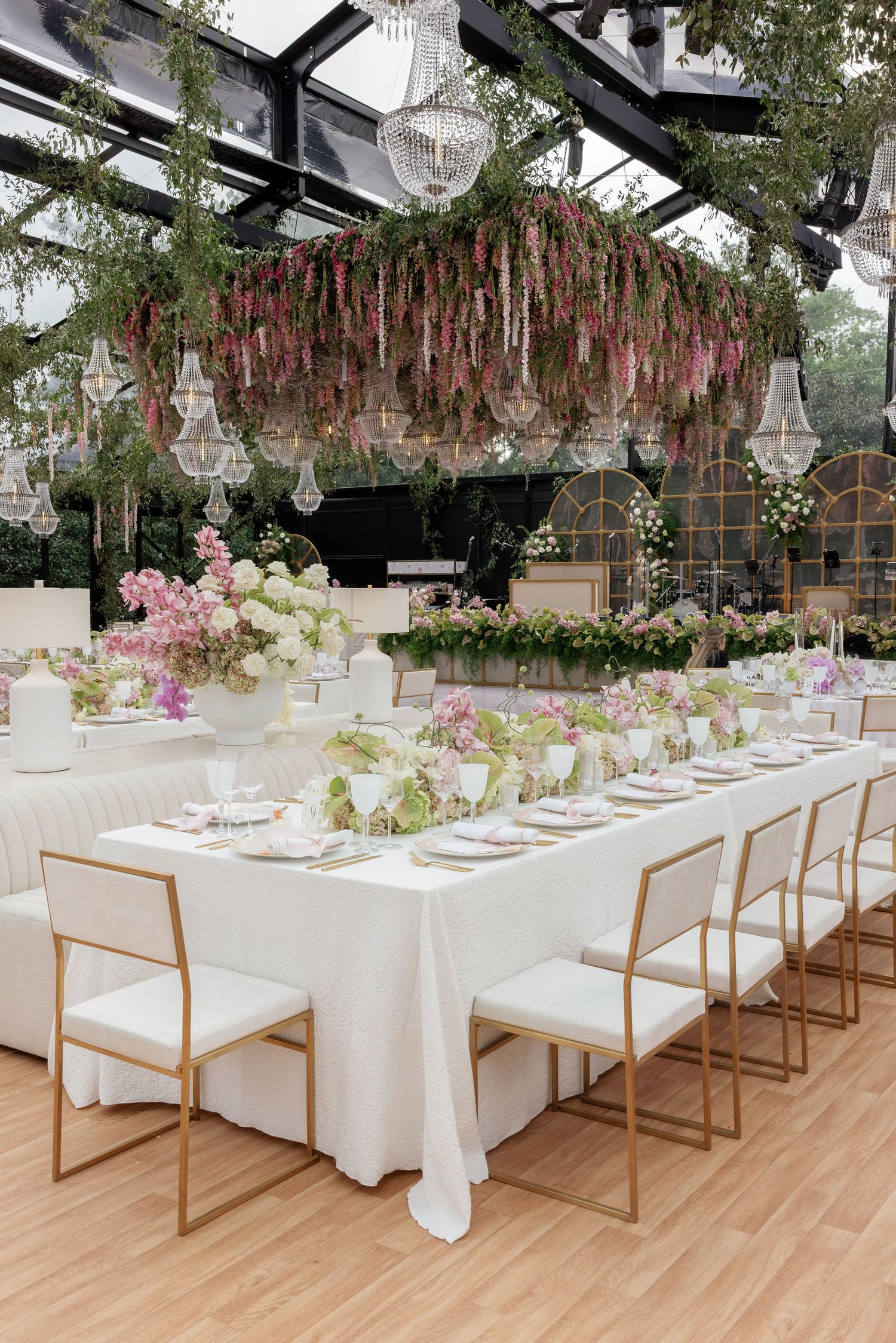 Elegant white and pink wedding reception setup under a greenhouse with hanging floral arrangements and modern decor.