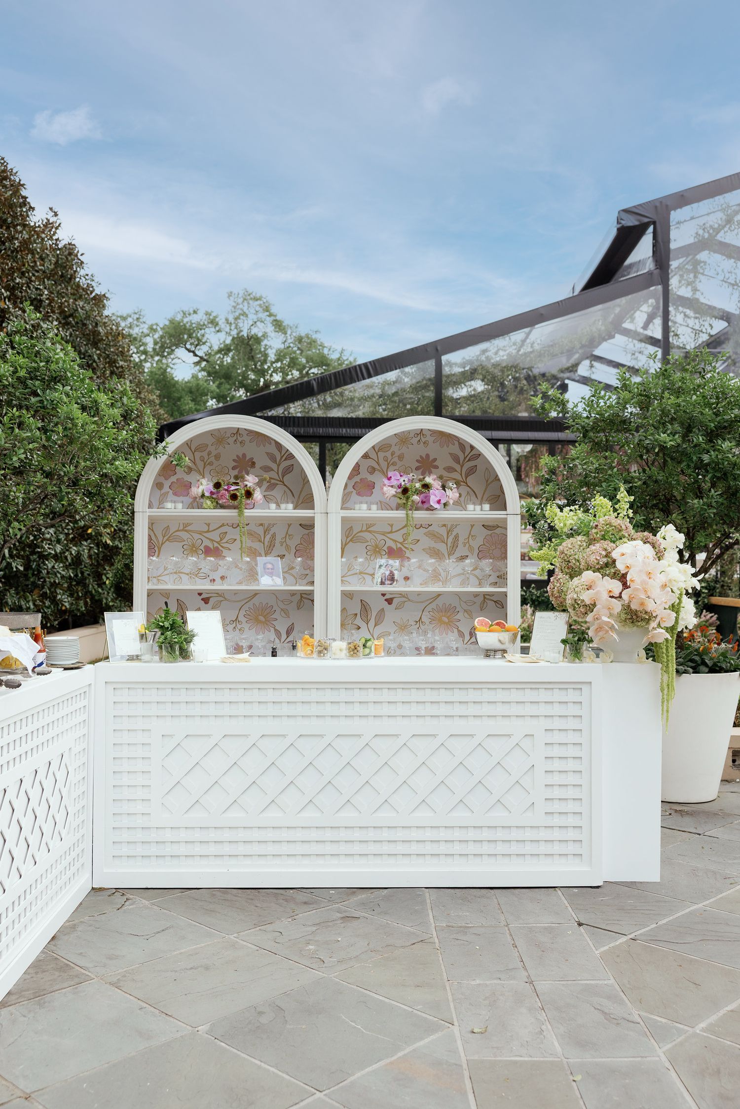 Elegant white display counter with arched shelving showcases soft pink floral arrangements at an outdoor garden venue.