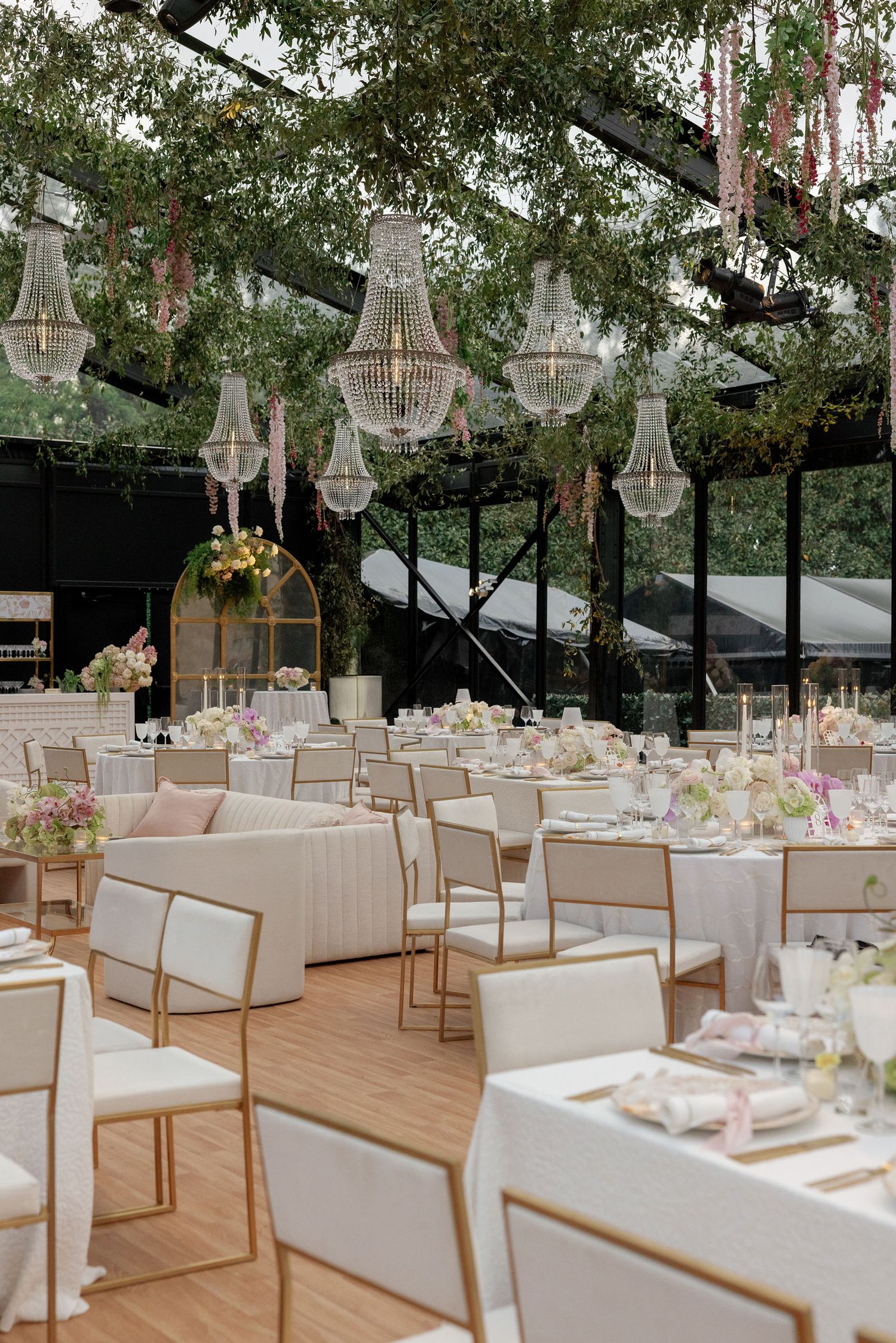 Elegant wedding reception setup in a clear tent with crystal chandeliers, wooden chairs and white tablecloths.