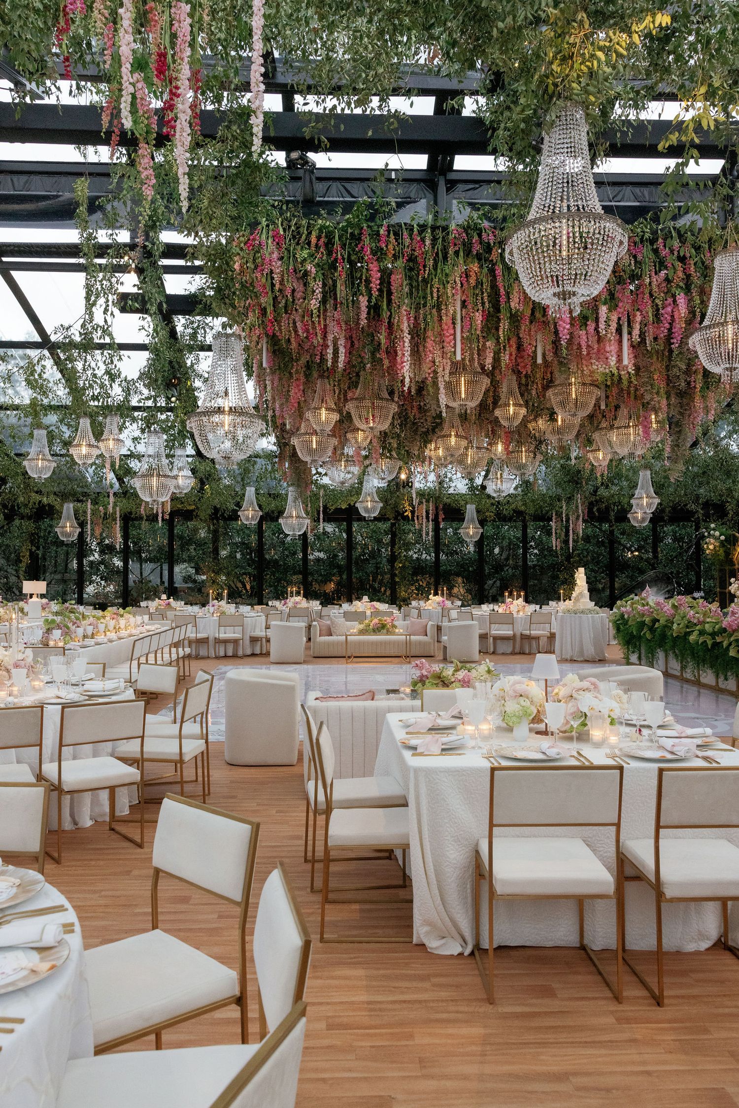 Elegant wedding reception venue with crystal chandeliers, white tables, and pink floral arrangements under a glass ceiling.