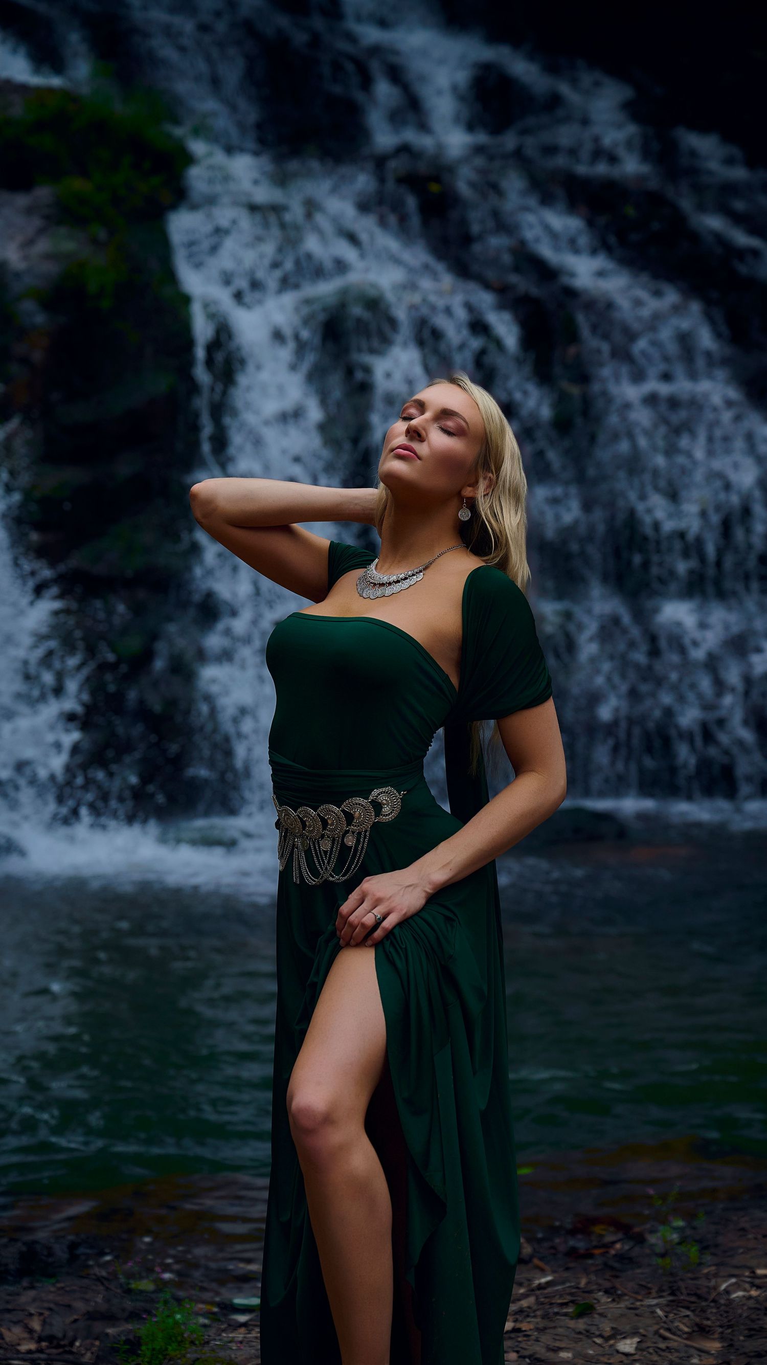 A person in an elegant dark green dress poses dramatically in front of a cascading waterfall.