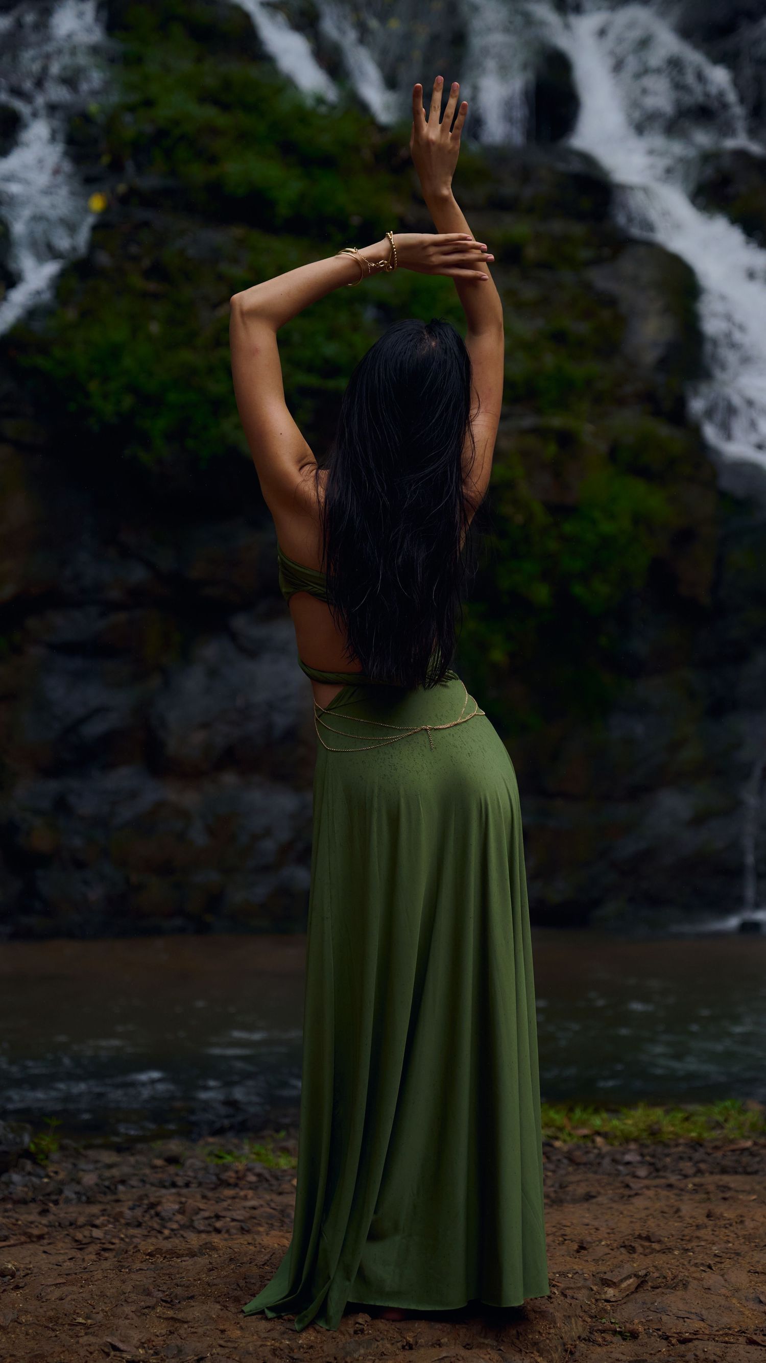 Person in olive green dress poses gracefully near a cascading waterfall and rocky shoreline.