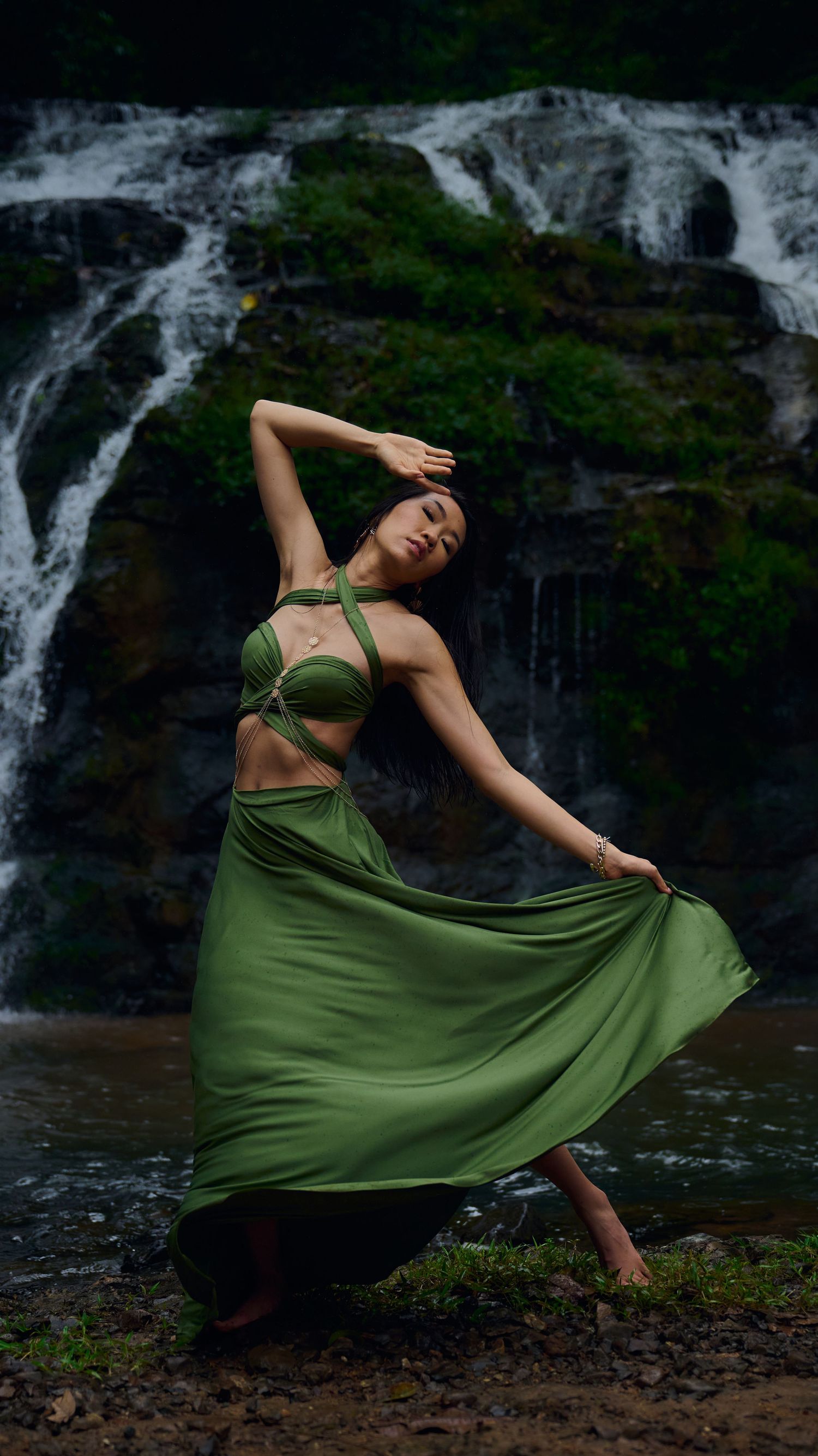 Graceful dancer in flowing green dress performs expressive movements against backdrop of cascading waterfall in nature.