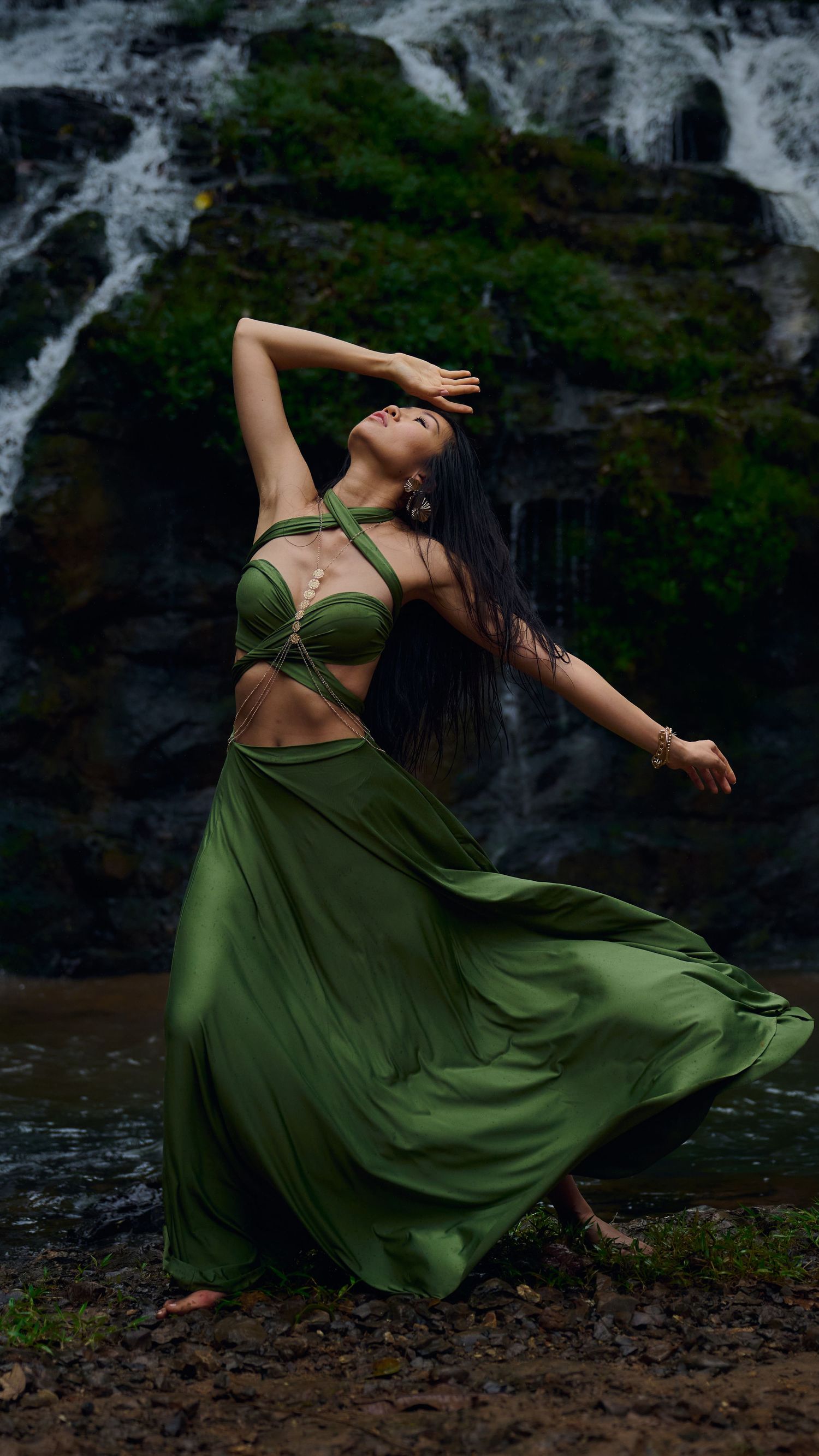 Dancer in flowing olive green dress poses gracefully against dramatic waterfall background in nature setting.