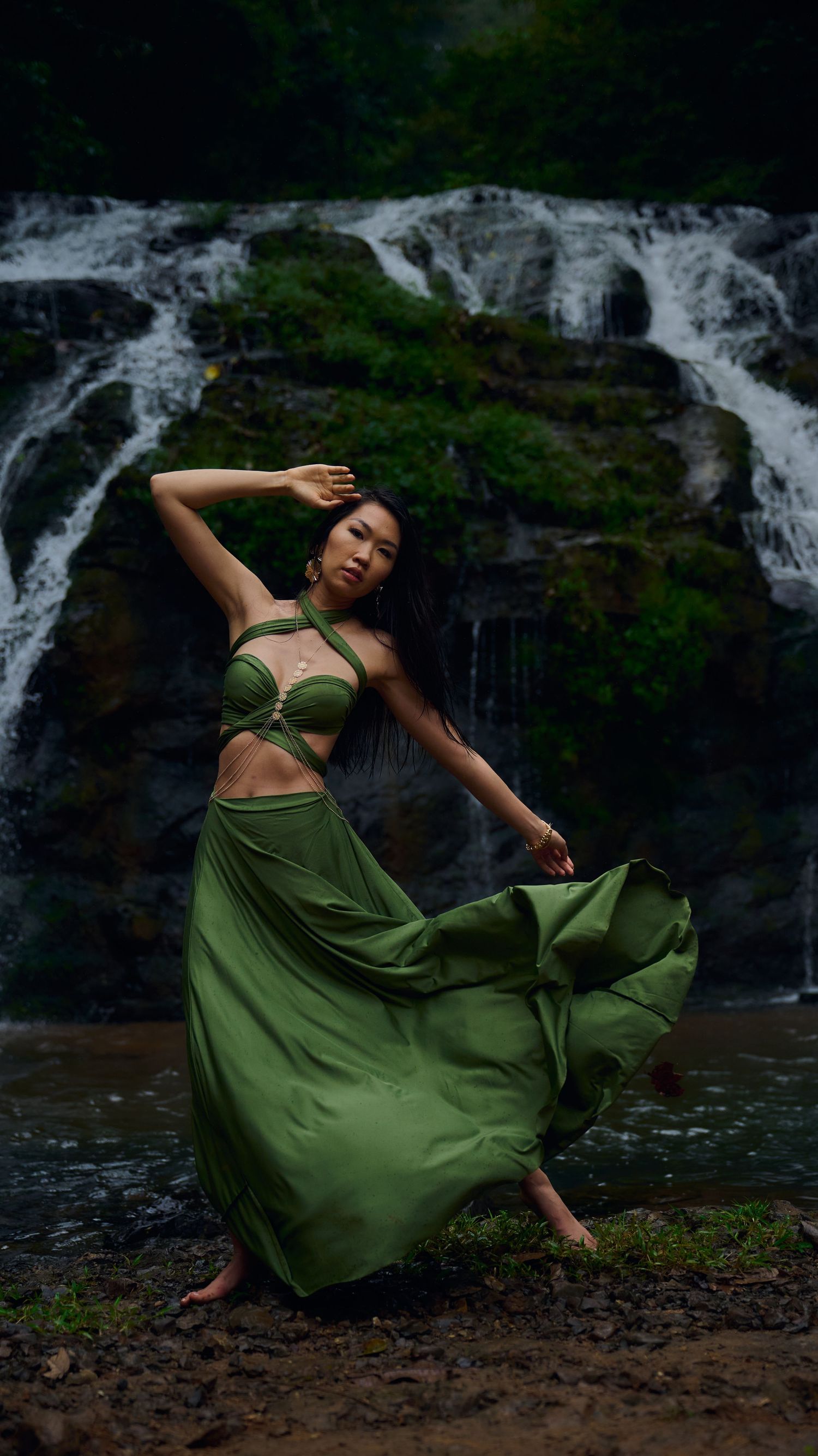 A dancer in a flowing olive green dress performs graceful movements against a scenic waterfall backdrop.