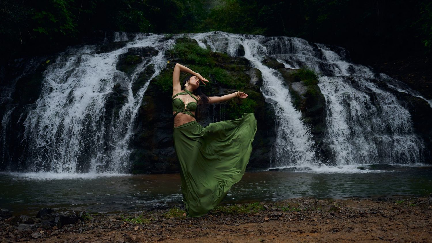 A dancer in a flowing green dress performs gracefully in front of a cascading waterfall in a forest setting.
