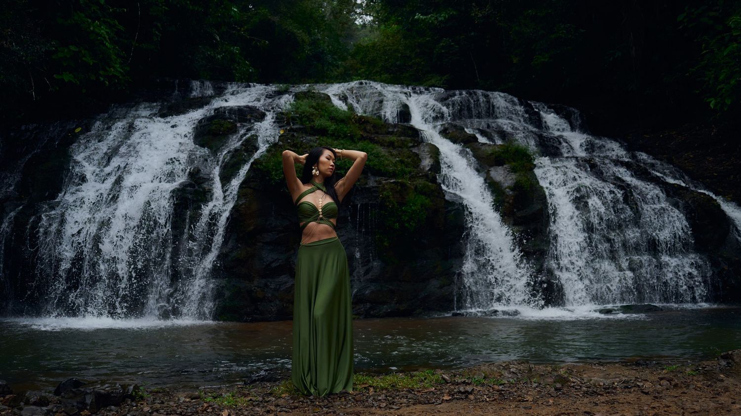 Person in flowing green dress stands with raised arms against a scenic cascading waterfall in a lush forest setting.