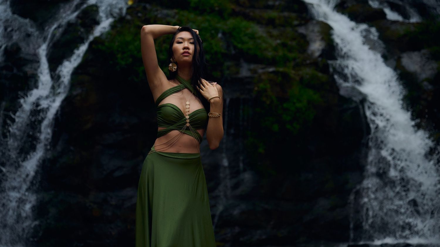 A person in a green outfit poses against a scenic double waterfall backdrop in a lush forest setting.