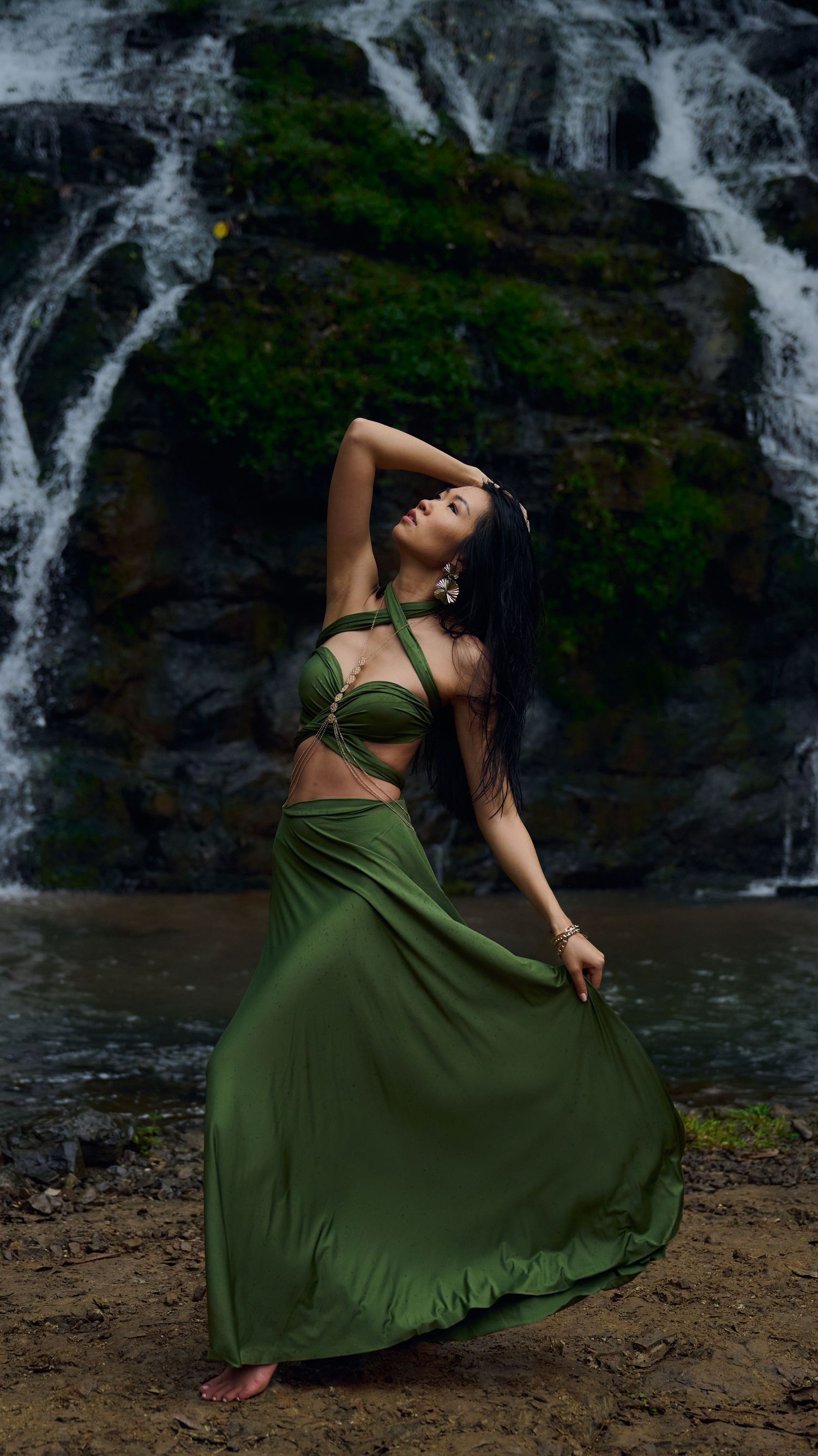 A graceful dance pose in an olive green outfit against a cascading waterfall backdrop in nature.