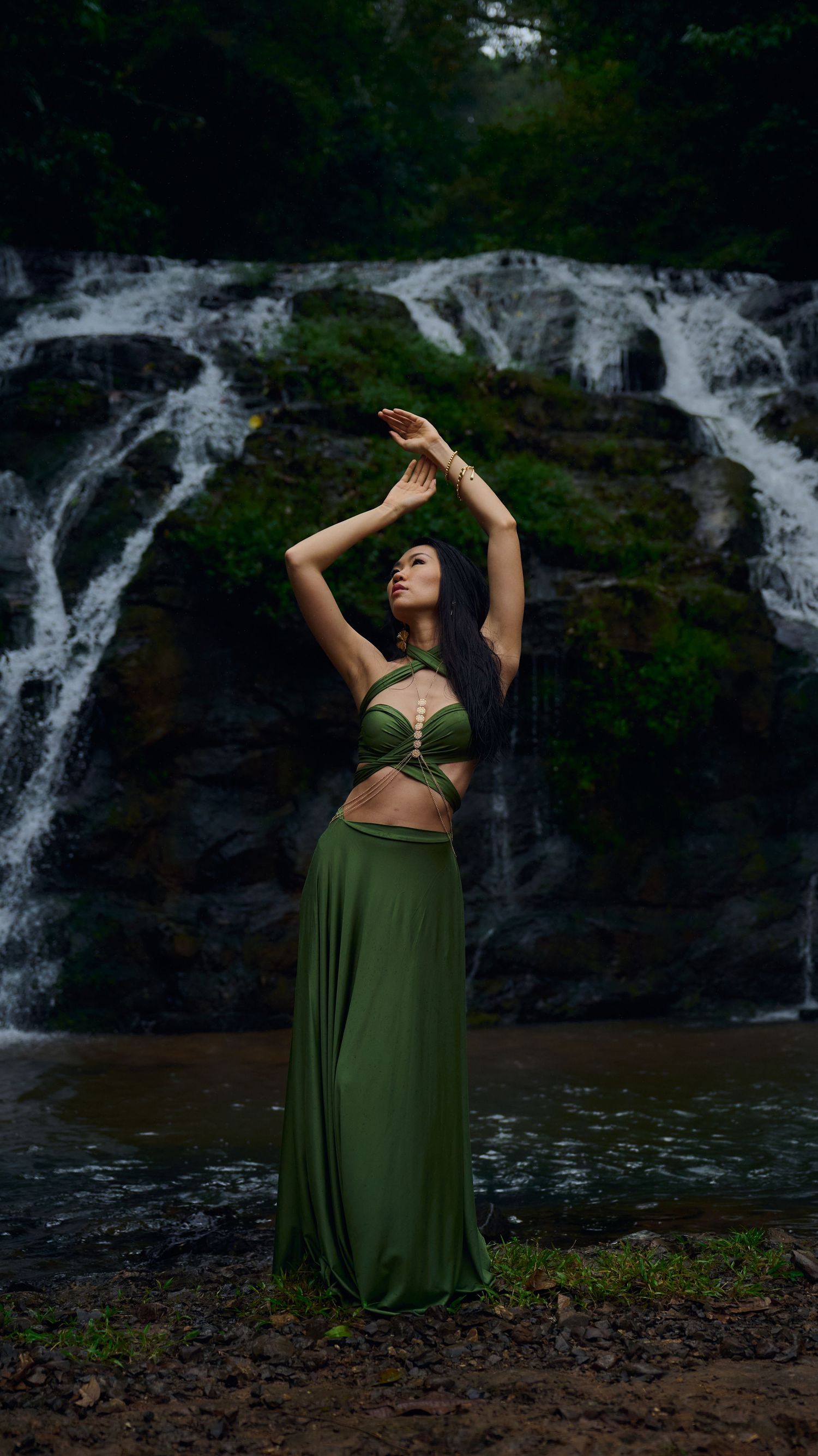 A dramatic dance pose performed in front of a scenic waterfall with arms raised gracefully skyward.