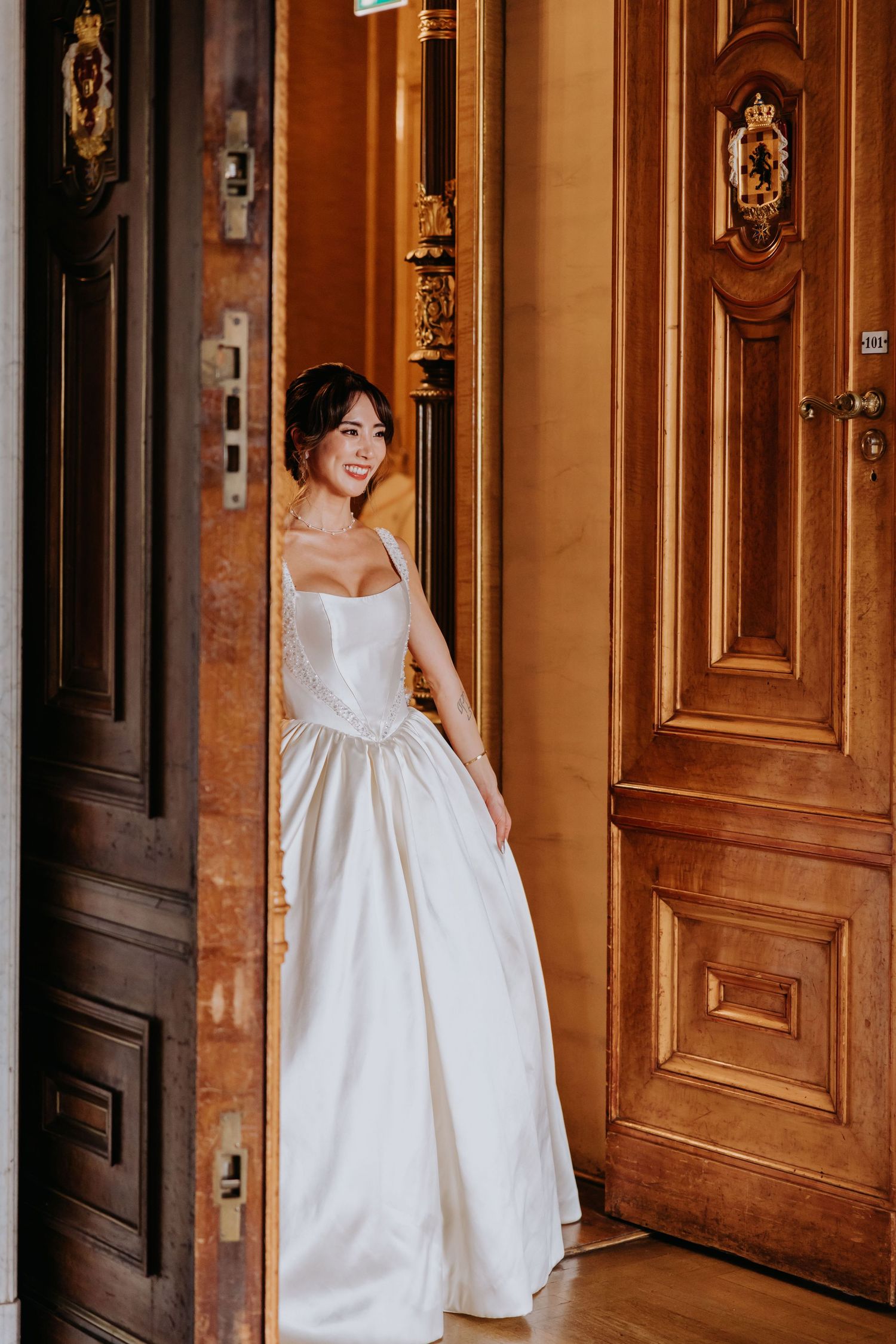 A person wearing a white empire waist dress poses in an elegant hallway with wooden doors and ornate trim details.