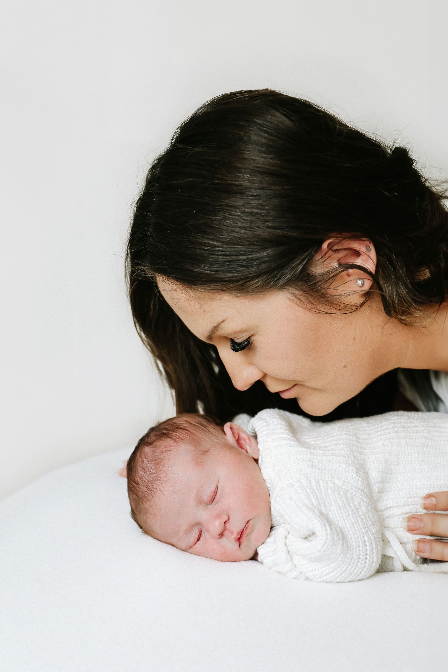 A tender moment between a mum and a sleeping newborn baby in white clothing on a light background, during newborn shoot in Nottingham