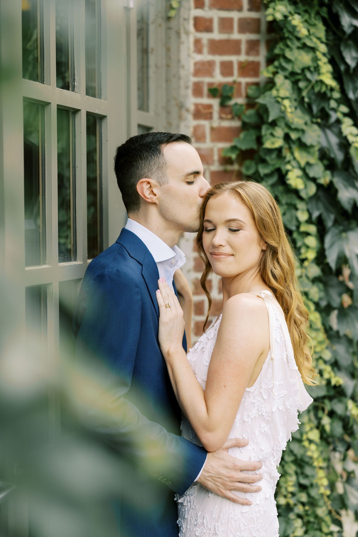 Couple embraces tenderly against brick wall with ivy in romantic outdoor setting.