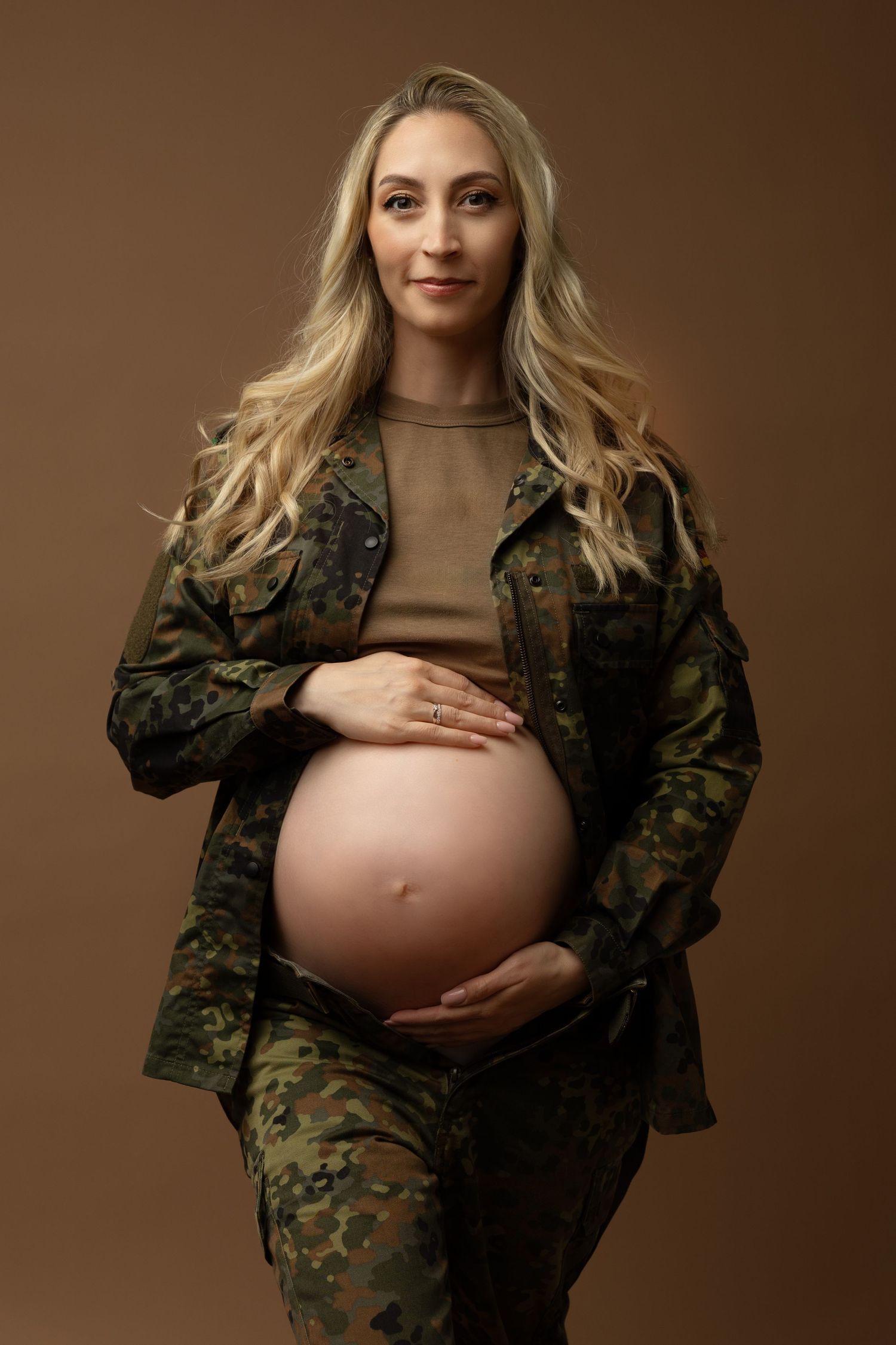 Maternity photo series showing pregnant woman in camouflage jacket against brown backdrop.