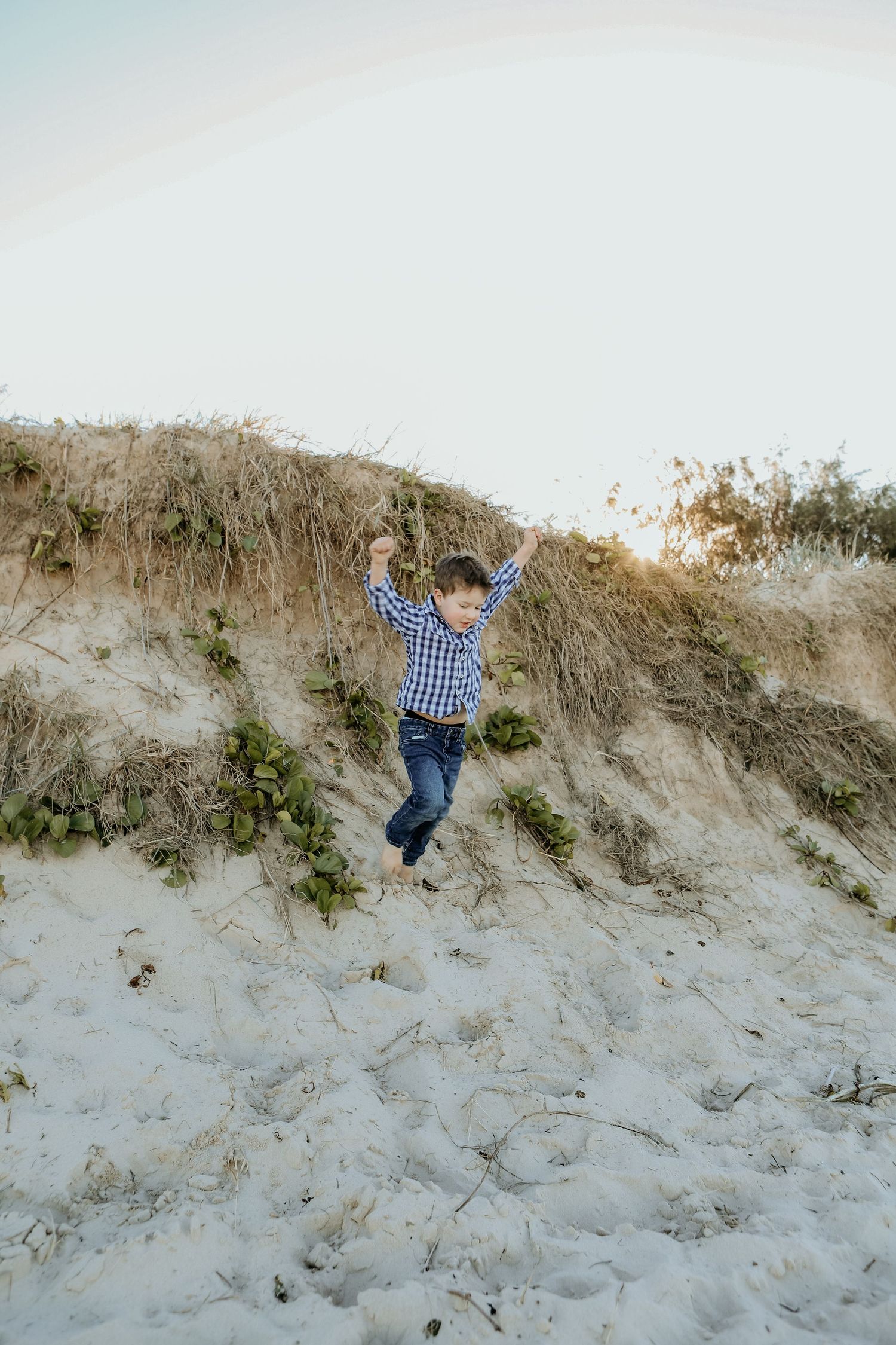 Young child in blue plaid shirt jumps and plays on sandy beach dunes on a cloudy day.