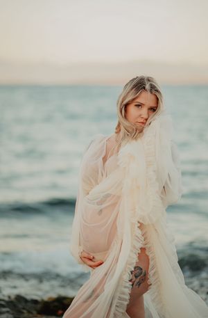 A person in a flowing cream dress poses along a misty beach at sunset with ethereal fabric billowing in the wind.