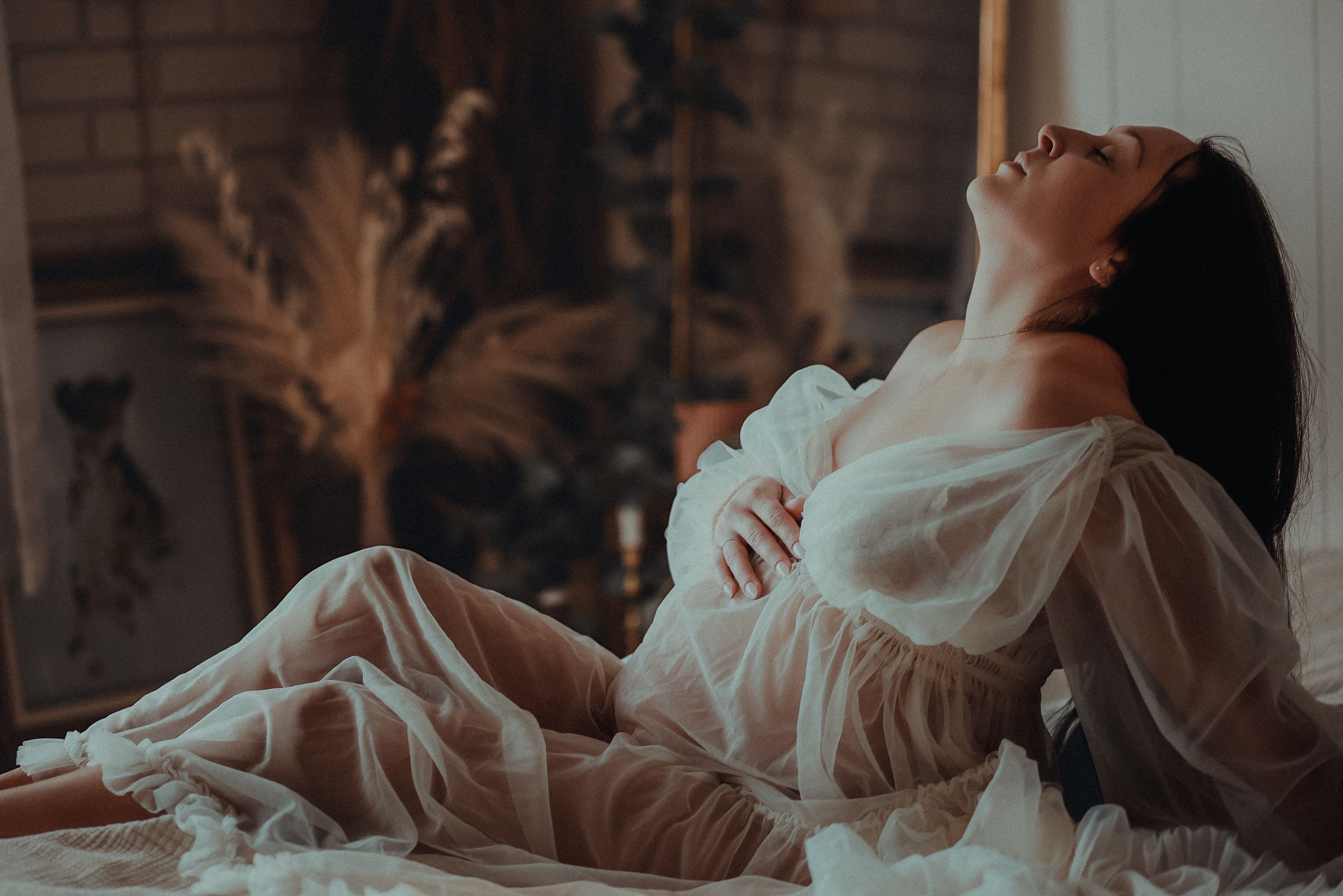 A woman in a flowing white dress reclines on a bed with soft lighting and pampas grass decor in the background.