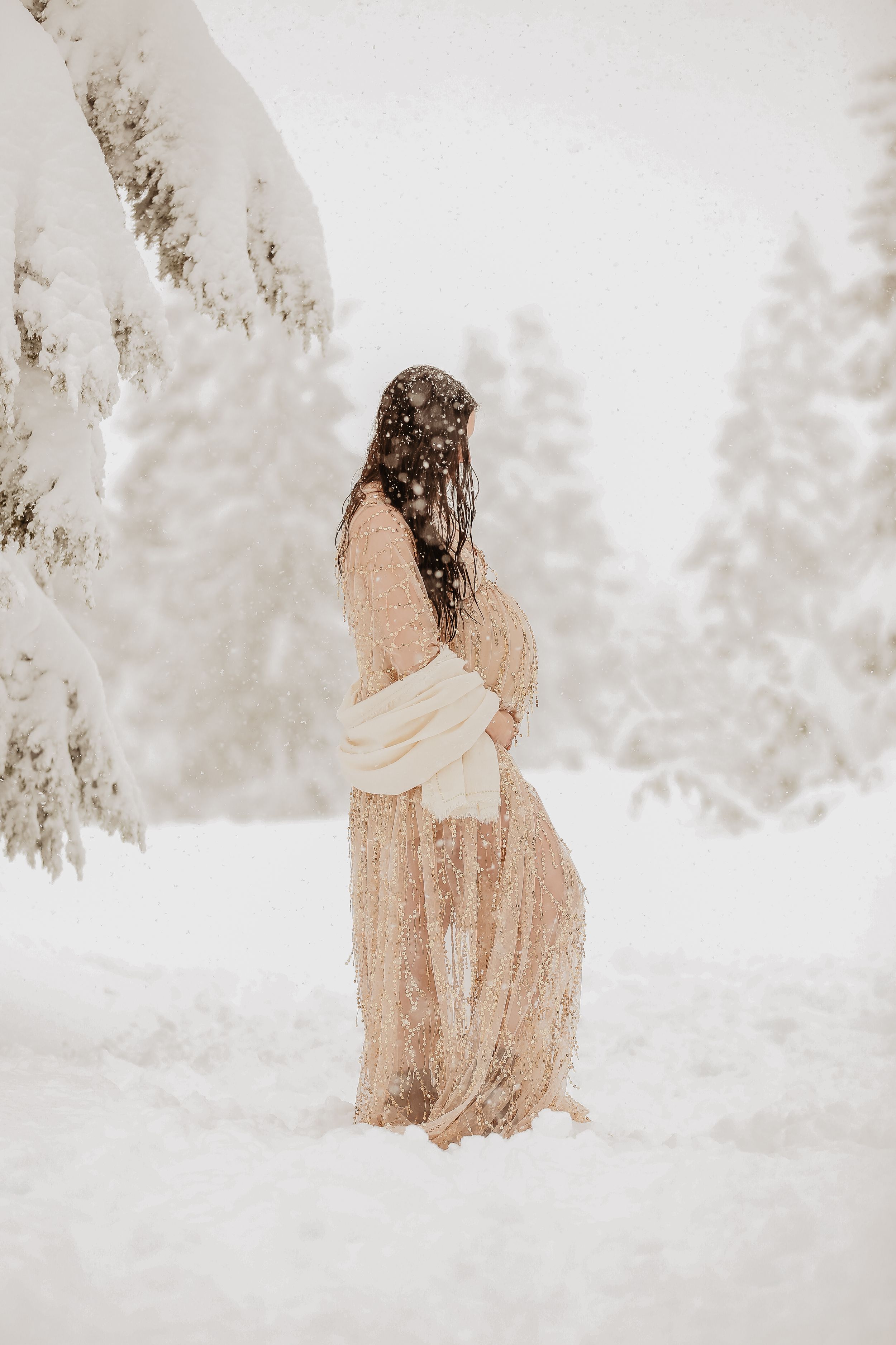 A figure in a flowing beige dress sits contemplatively in fresh white snow with misty winter forest in the background.