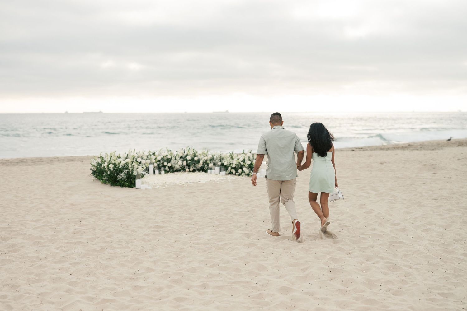 Two people walk along a serene beach at sunset beside an elegant floral arrangement on the sand.